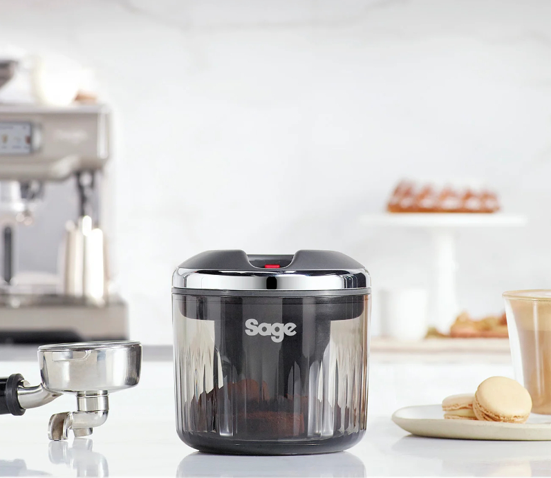 Eine Kaffeemühle mit einem transparenten Behälter, gefüllt mit Kaffeepulver. Eine Kaffeemaschine im Hintergrund.