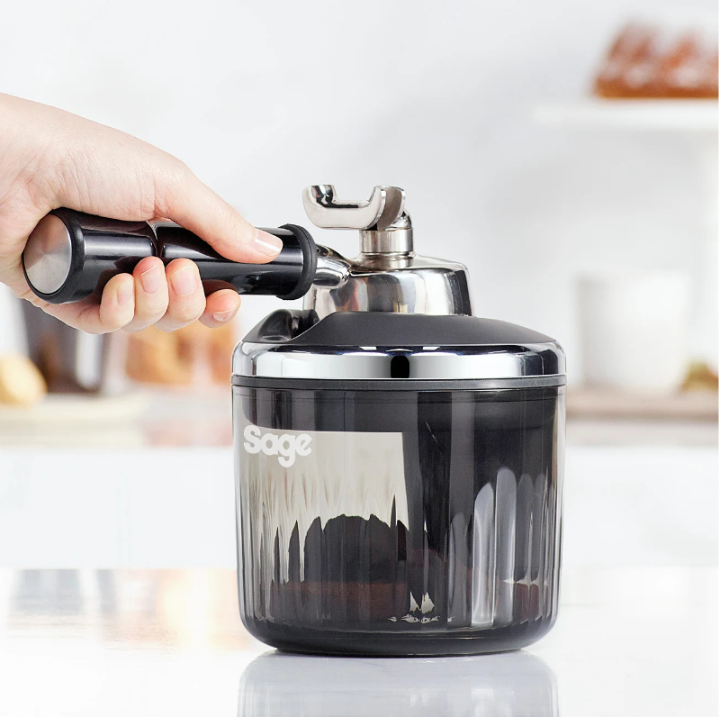 Hand hält Griff der Sage Kaffeemaschine. Schwarzes und silbernes Gerät auf weißer Oberfläche. Verschwommener Hintergrund mit Essen.