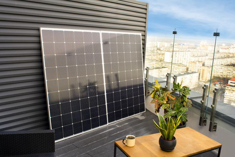 Zwei Solarmodule lehnen an einer grauen Wand, mit Pflanzen, Tisch und Stadt im Hintergrund.