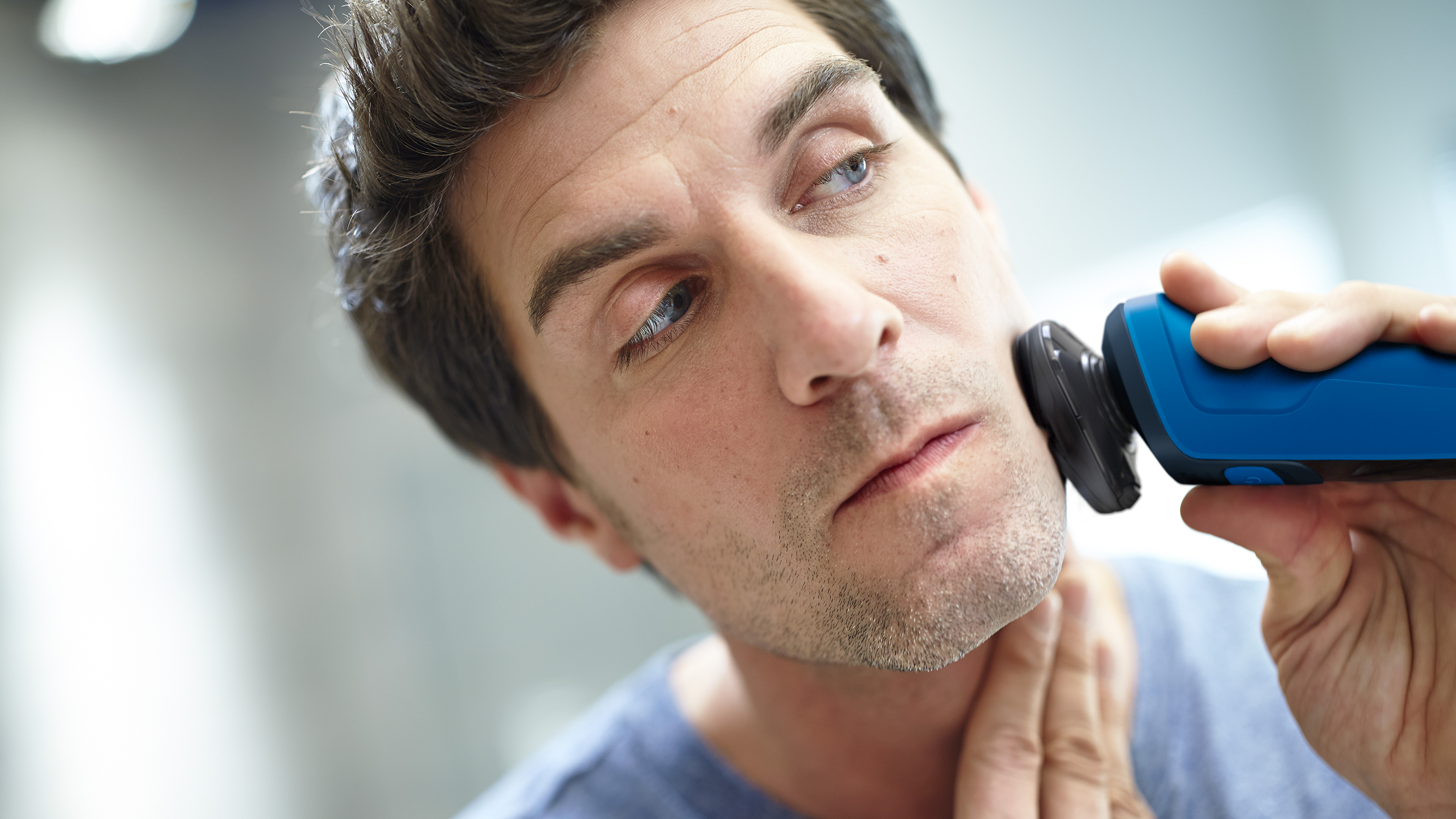 Man shaving his face with a blue and black electric shaver. Close-up, he is looking in the mirror.
