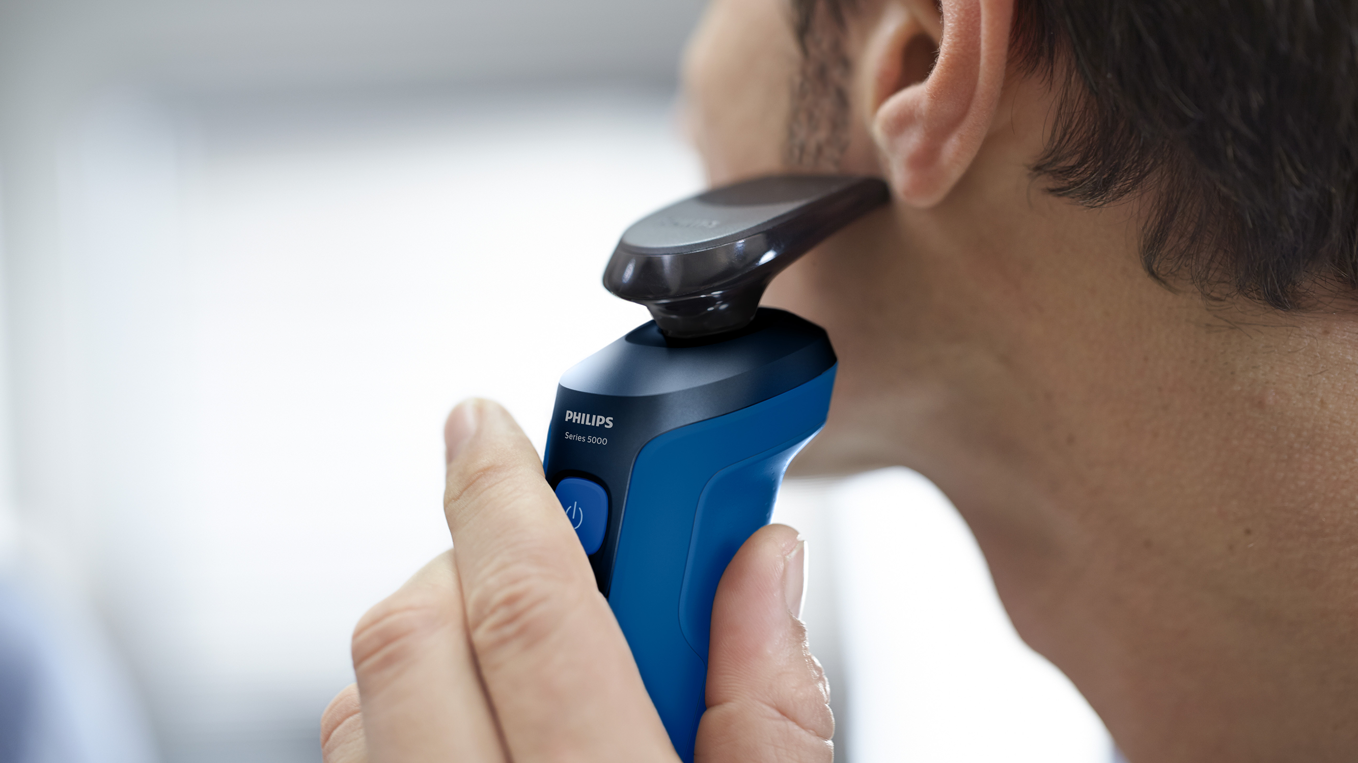 Close-up of a man shaving with a blue and black Philips electric razor. The model is Series 5000.