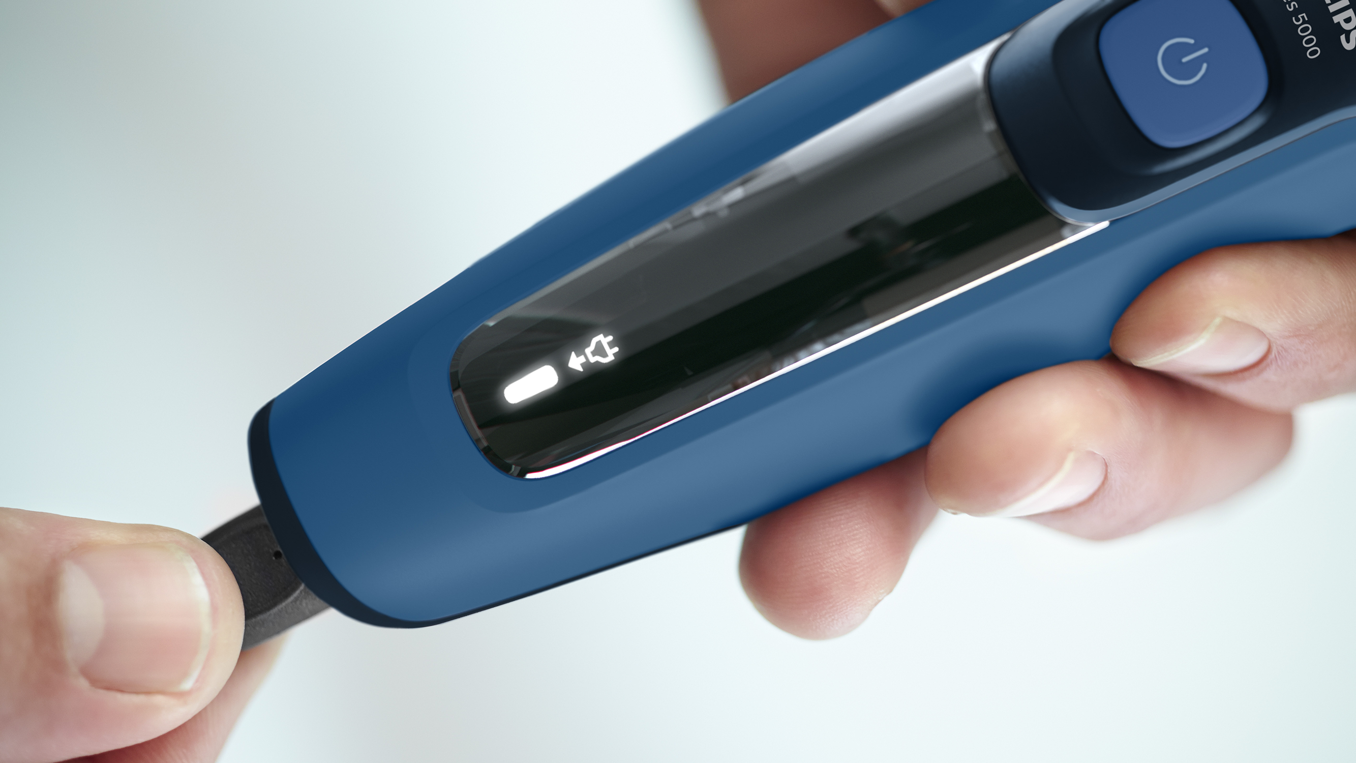 A blue electric shaver is held, a charging cable being plugged in. A light shows the charging status.