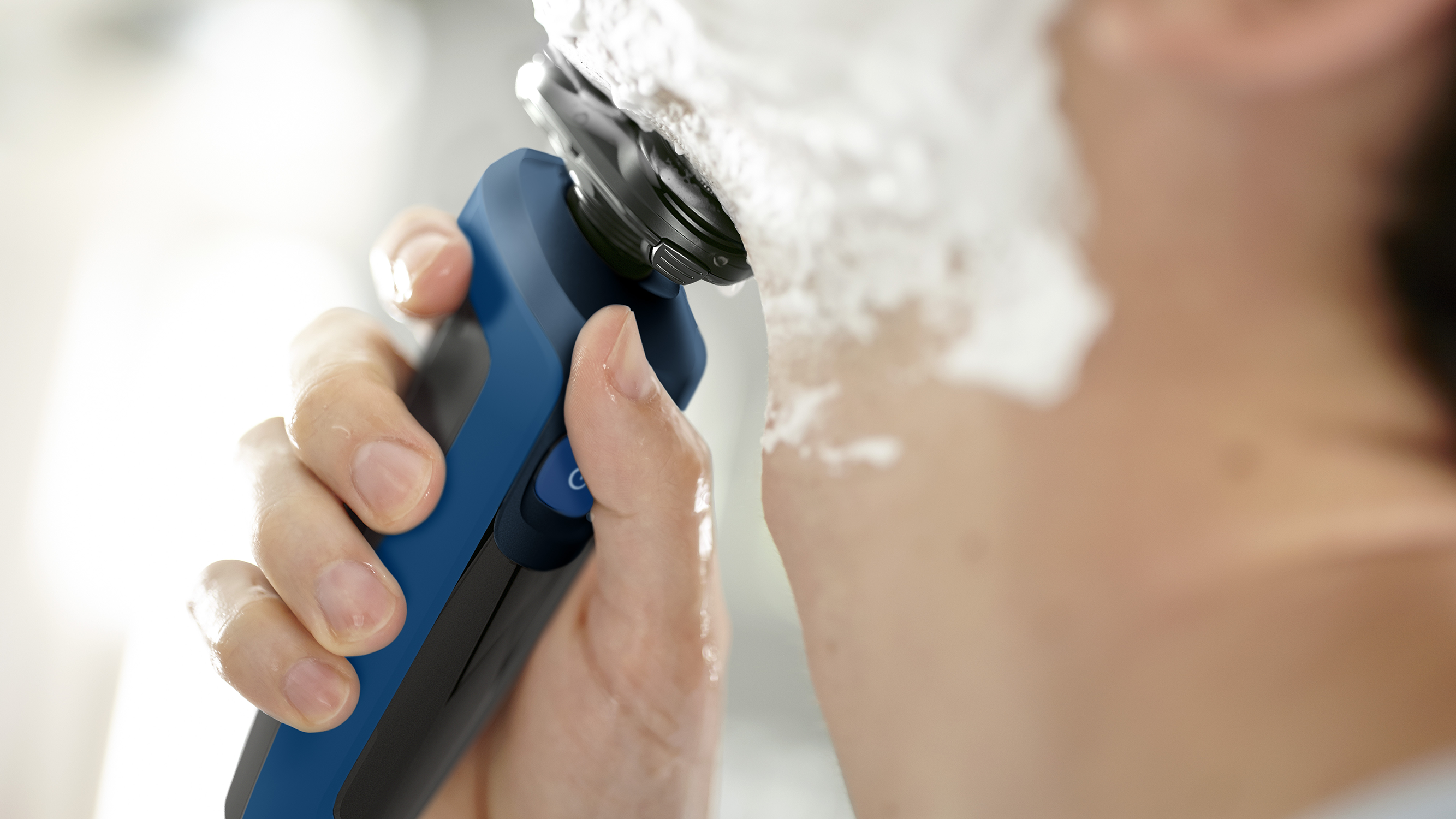 A man shaves his face with a blue and black electric shaver. Foam is visible.