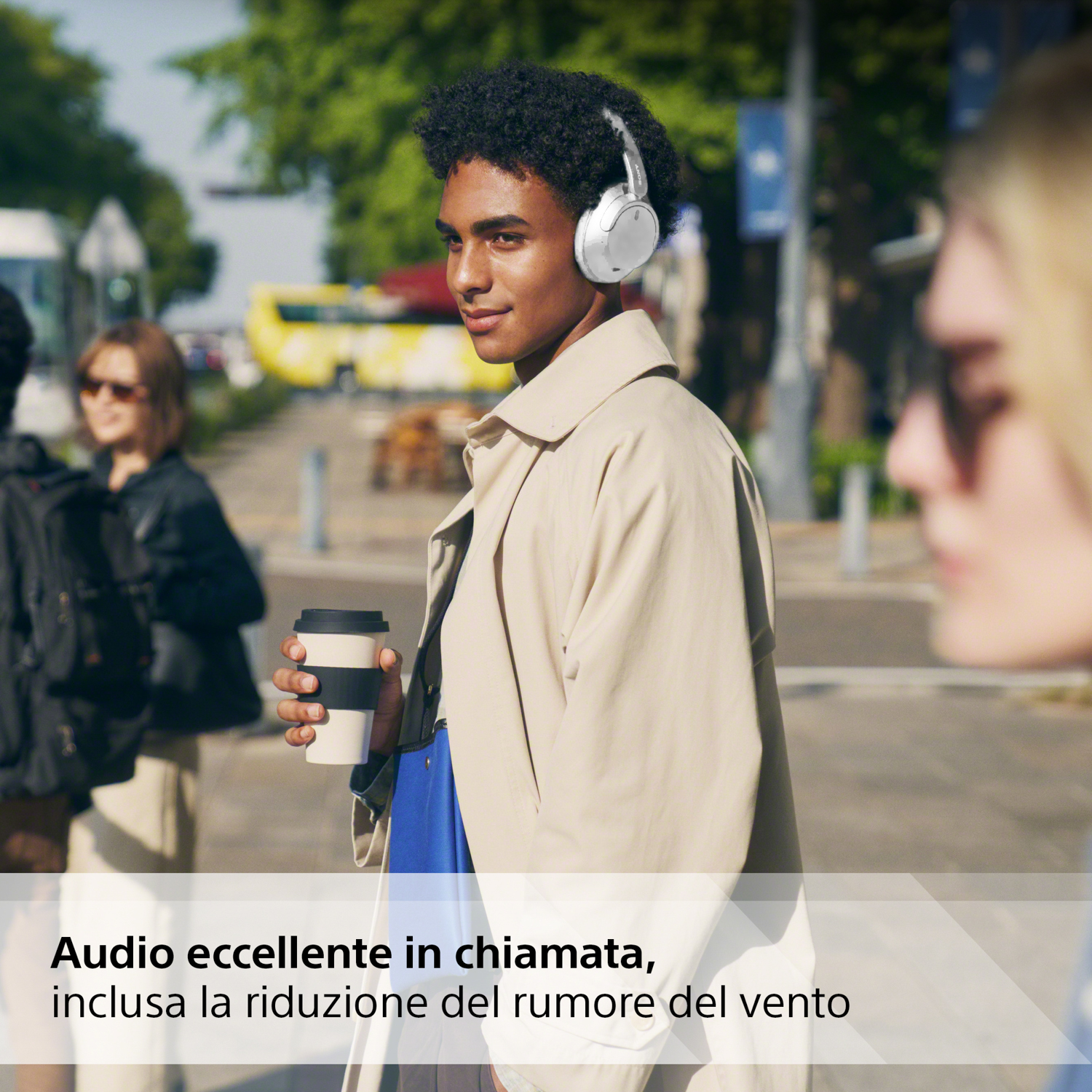 Un uomo con un cappotto color camoscio e cuffie bianche cammina per la strada, con un caffè. Altre persone sono presenti.