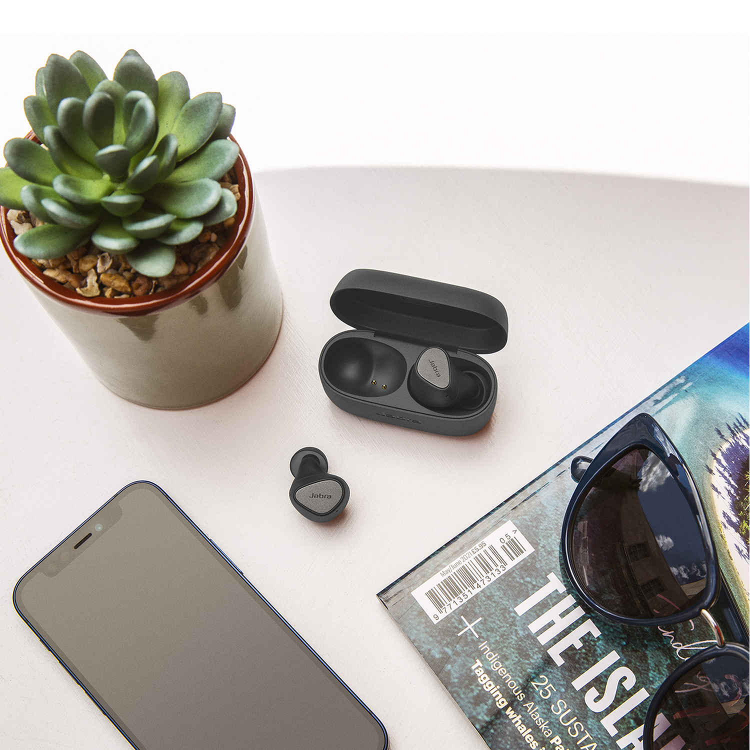 Oordopjes in een oplaadcase, met een telefoon, tijdschrift en zonnebril. Wit oppervlak.