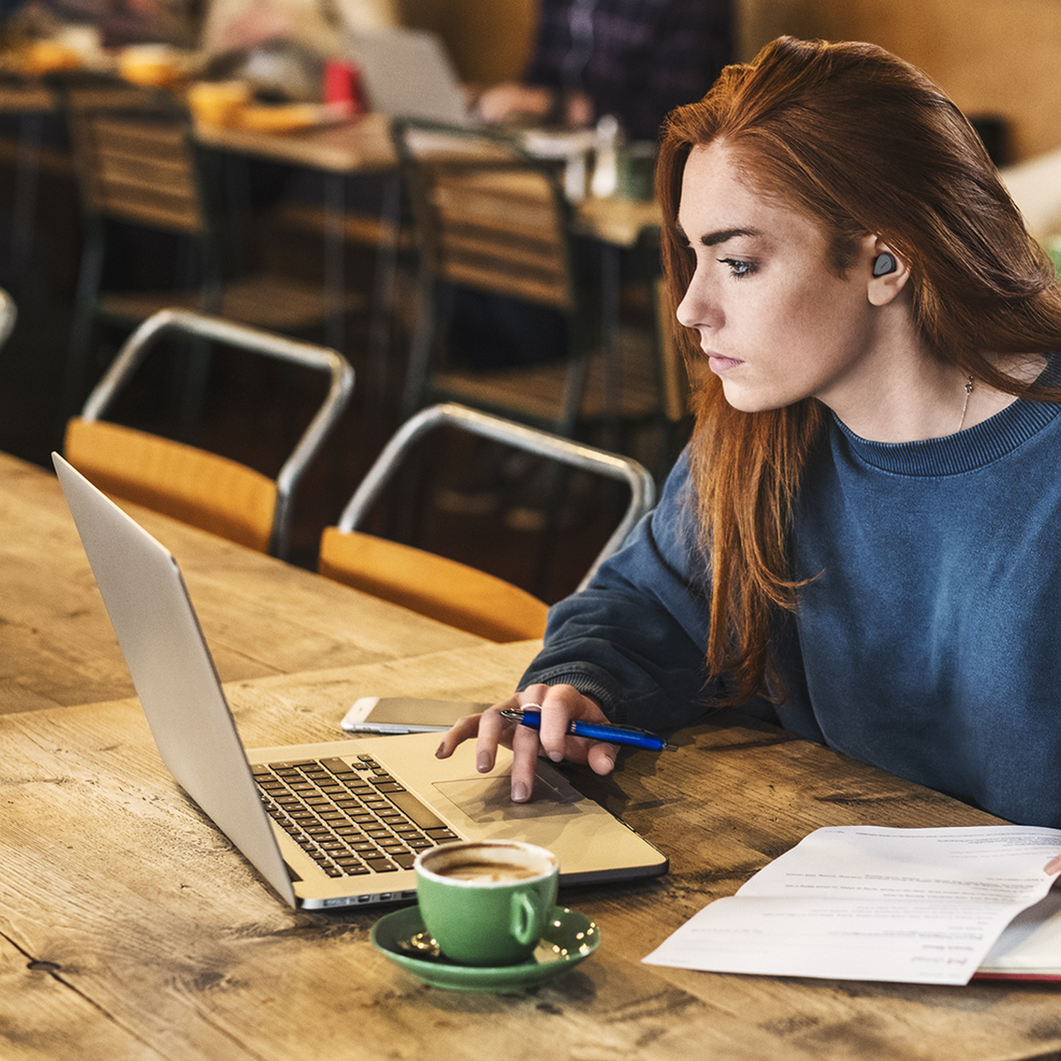 Vrouw met rood haar die een laptop gebruikt in een koffiebar. Ze heeft oordopjes in en kijkt geconcentreerd.