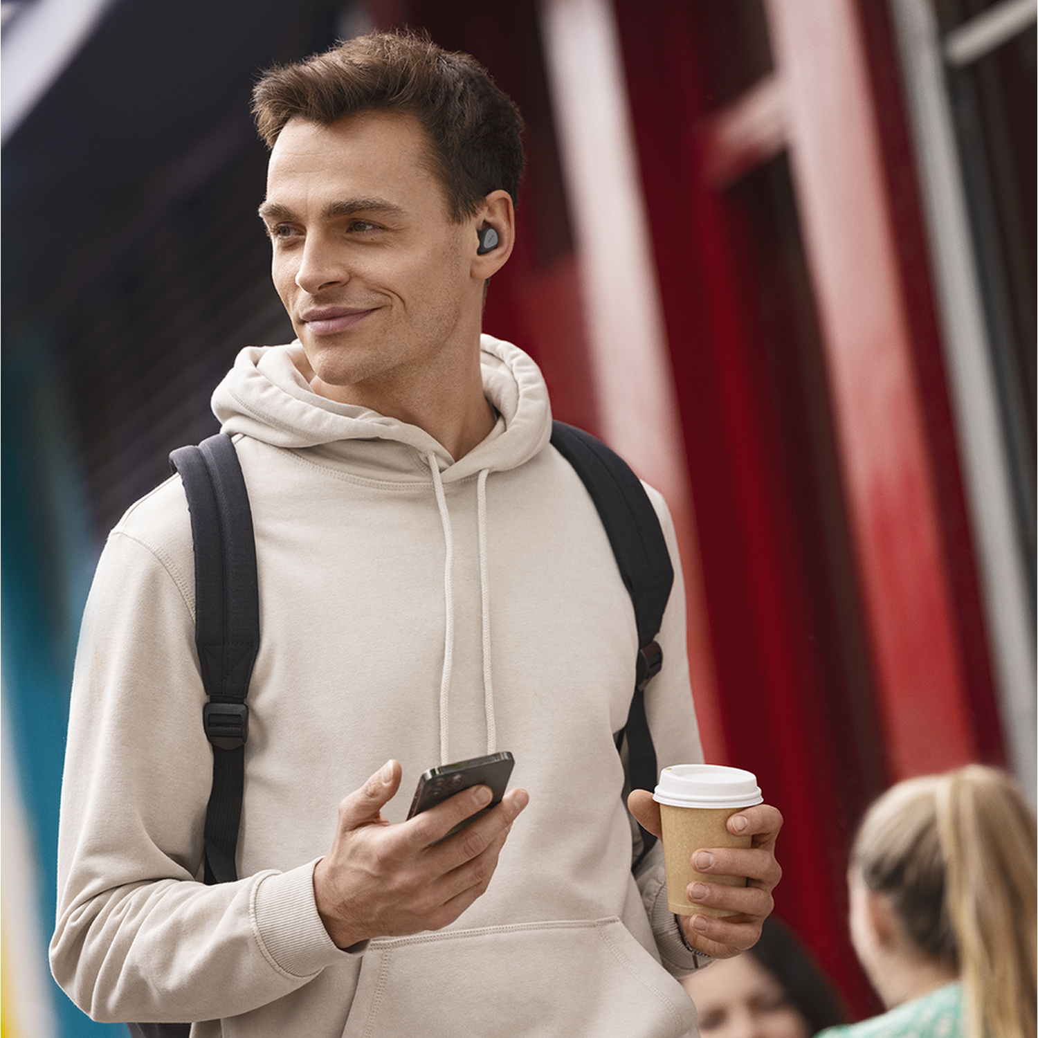 Man met oordopjes, telefoon en koffie, lachend buiten.