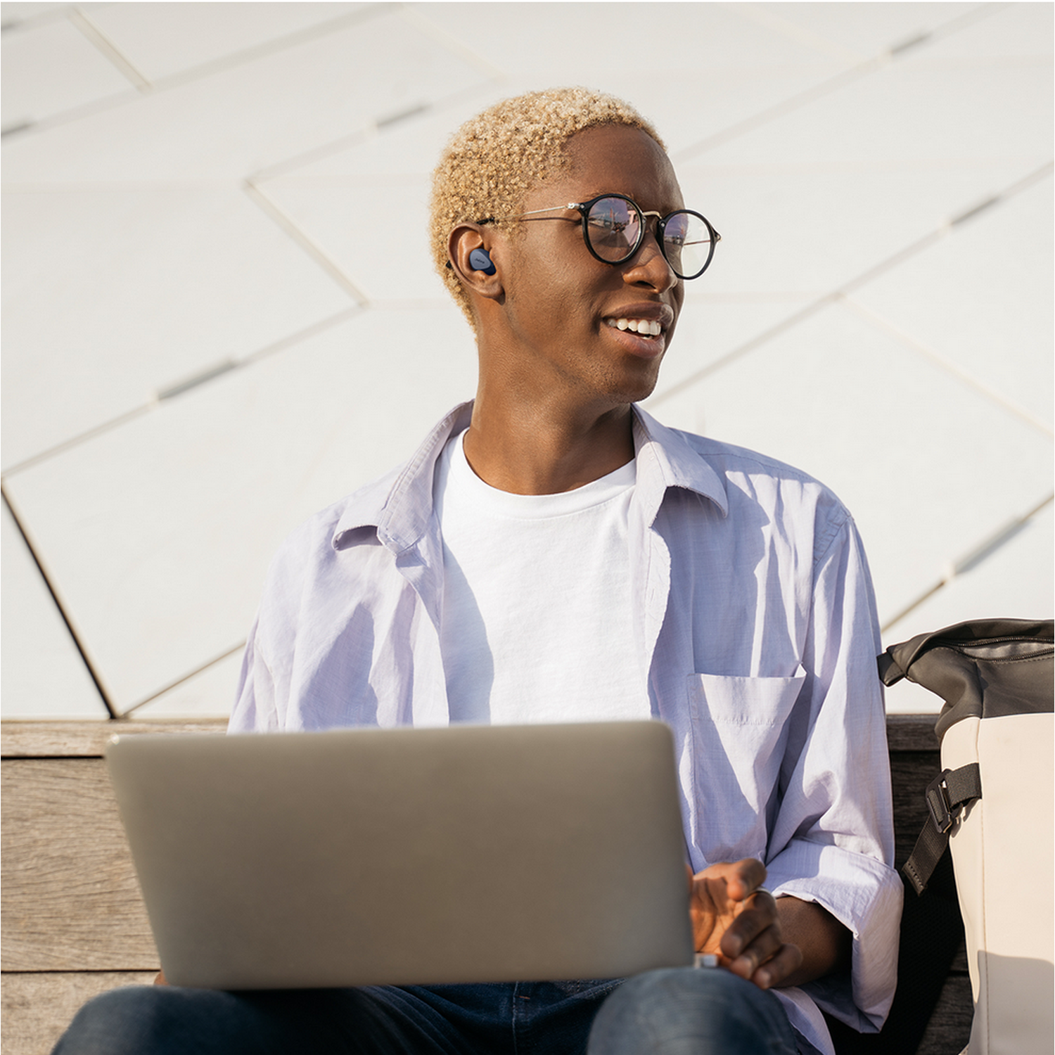 Een jonge man met blond haar en een bril gebruikt een laptop. Hij is buiten en glimlacht.