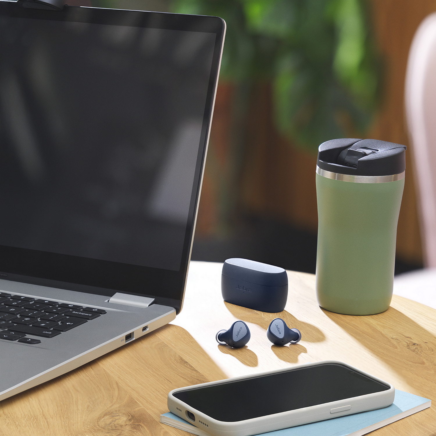 Een laptop, een telefoon, oordopjes en een koffiemok staan op een tafel.