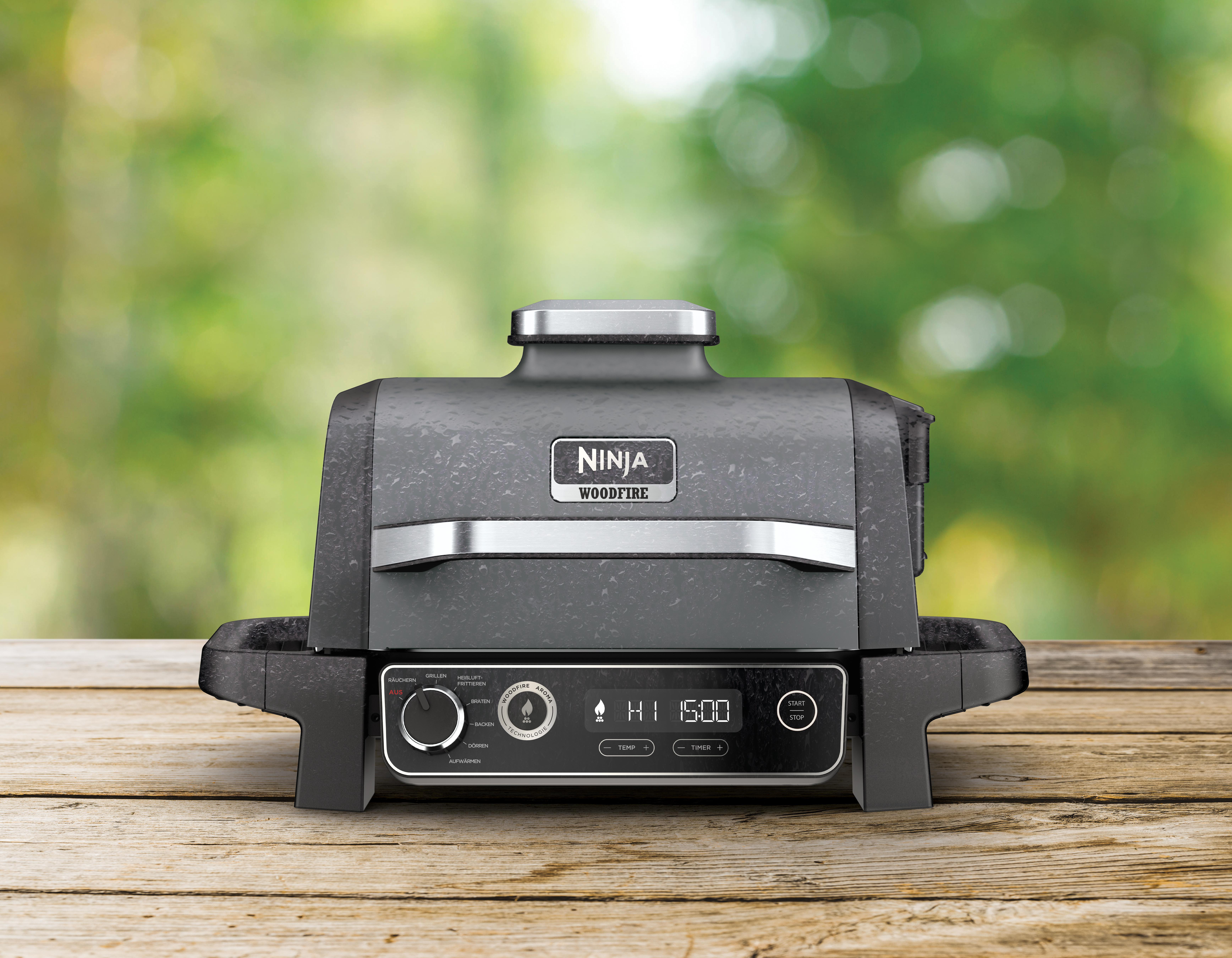 Ein schwarzer Ninja Woodfire Grill auf einem Holztisch, mit verschwommenem grünem Hintergrund.