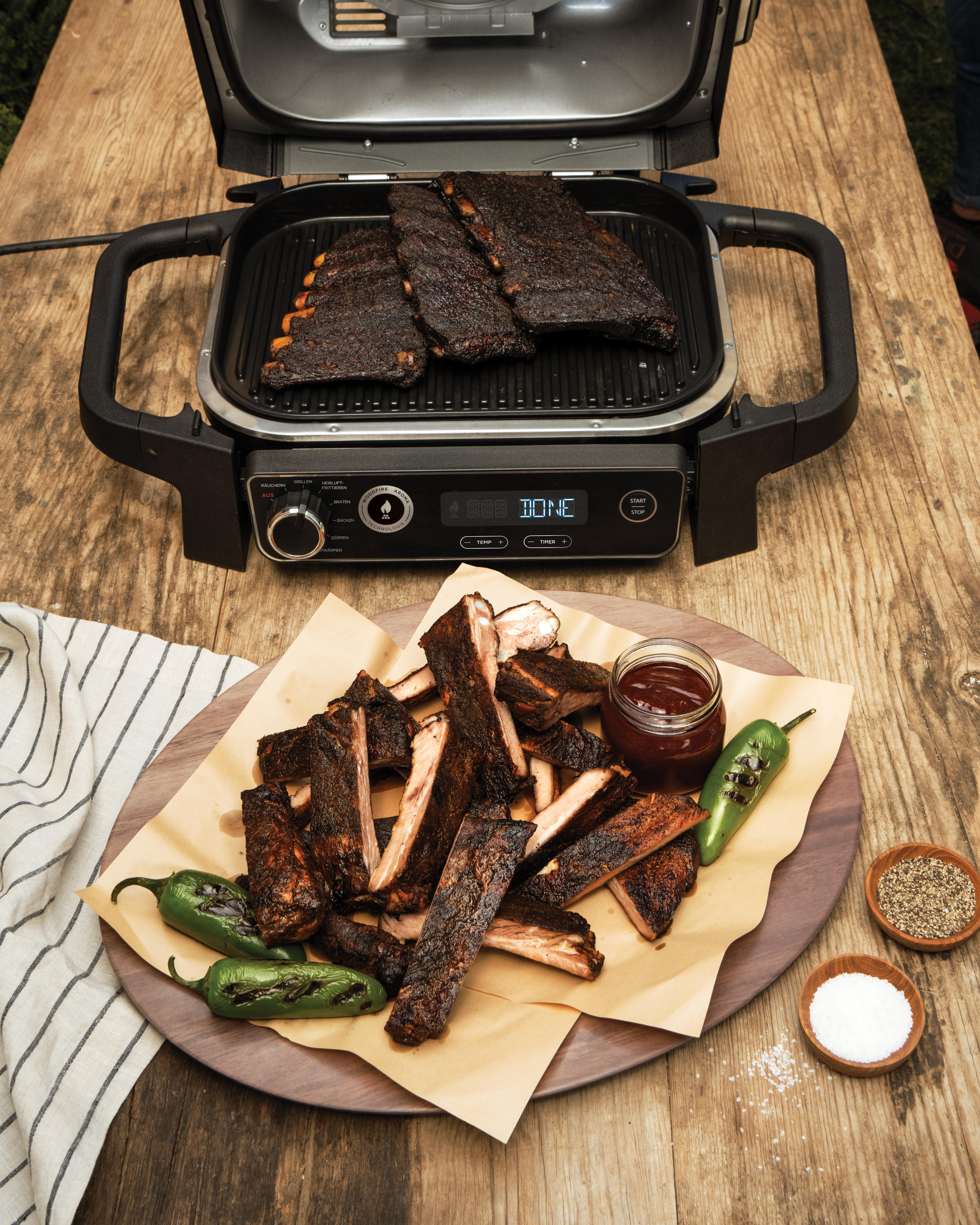 Gegrillte Rippchen liegen auf einem Grill. Geschnittene Rippchen, ein Glas Soße und Paprika sind auf einem Holzbrett angeordnet.