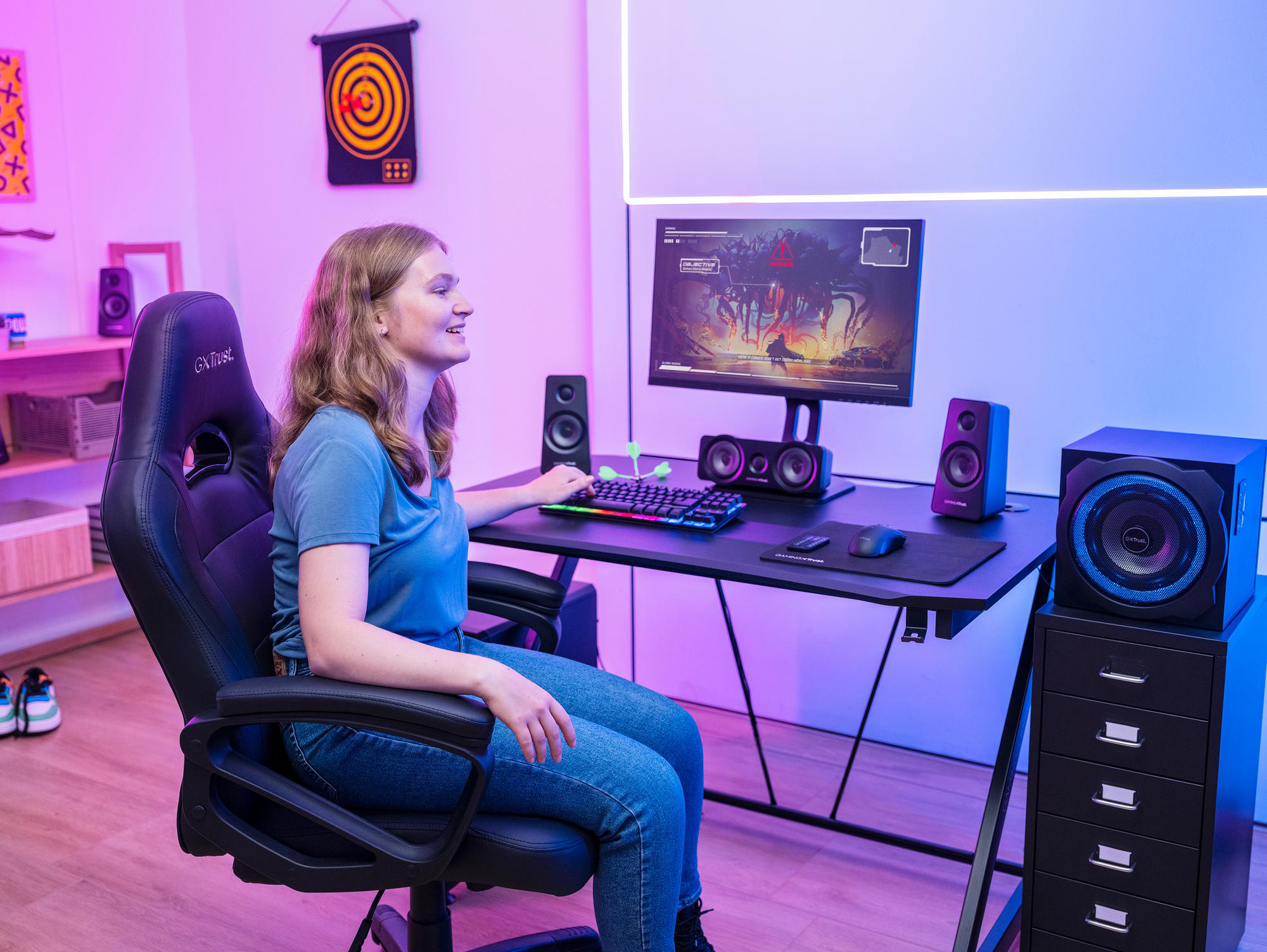 Eine junge Frau sitzt in einem Gaming-Stuhl vor einem beleuchteten Schreibtisch mit Monitor, Lautsprechern und PC-Spielen.