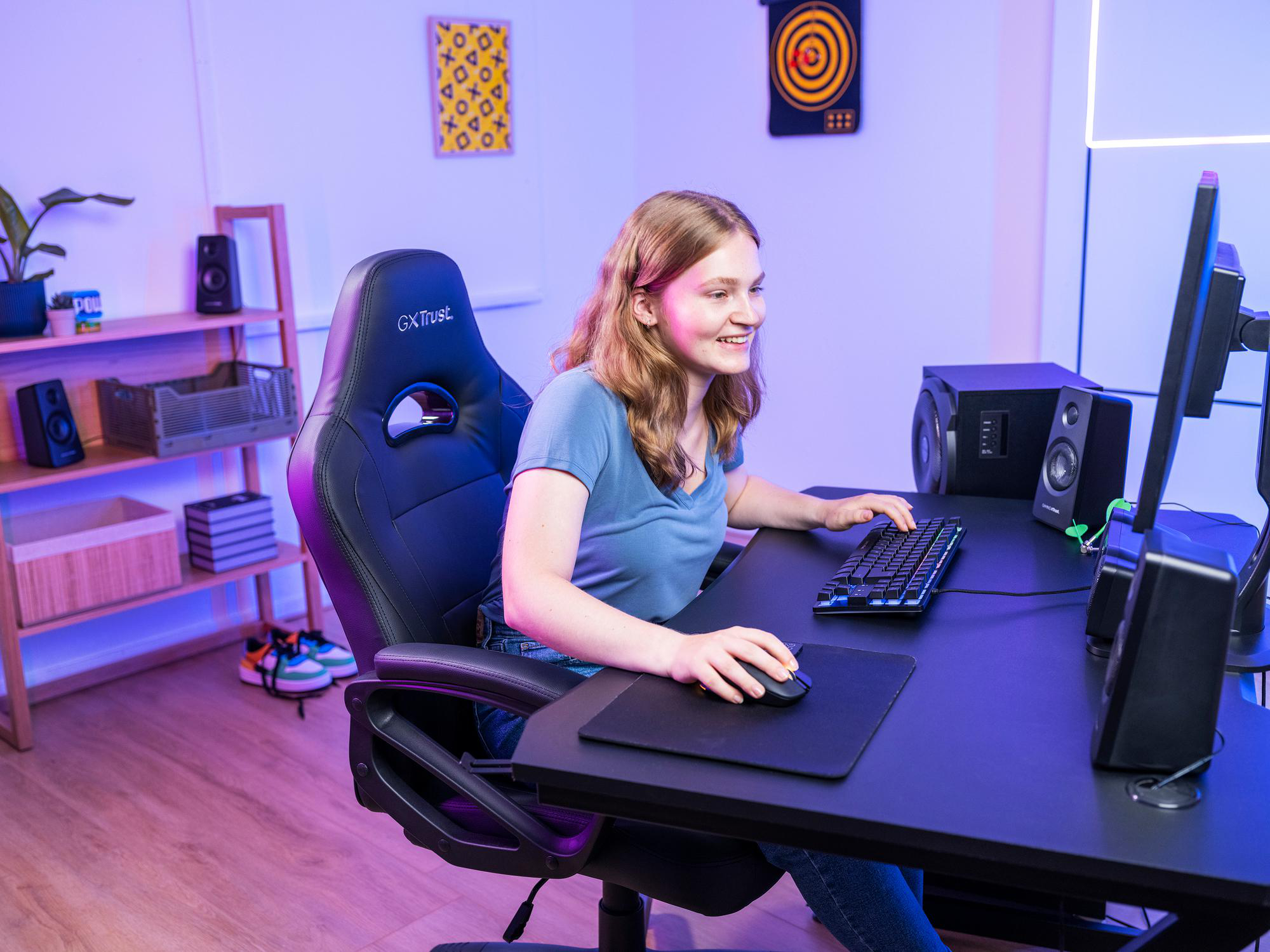 Eine junge Frau lächelt, während sie in einem beleuchteten Raum an einem Schreibtisch sitzt und Computerspiele spielt.