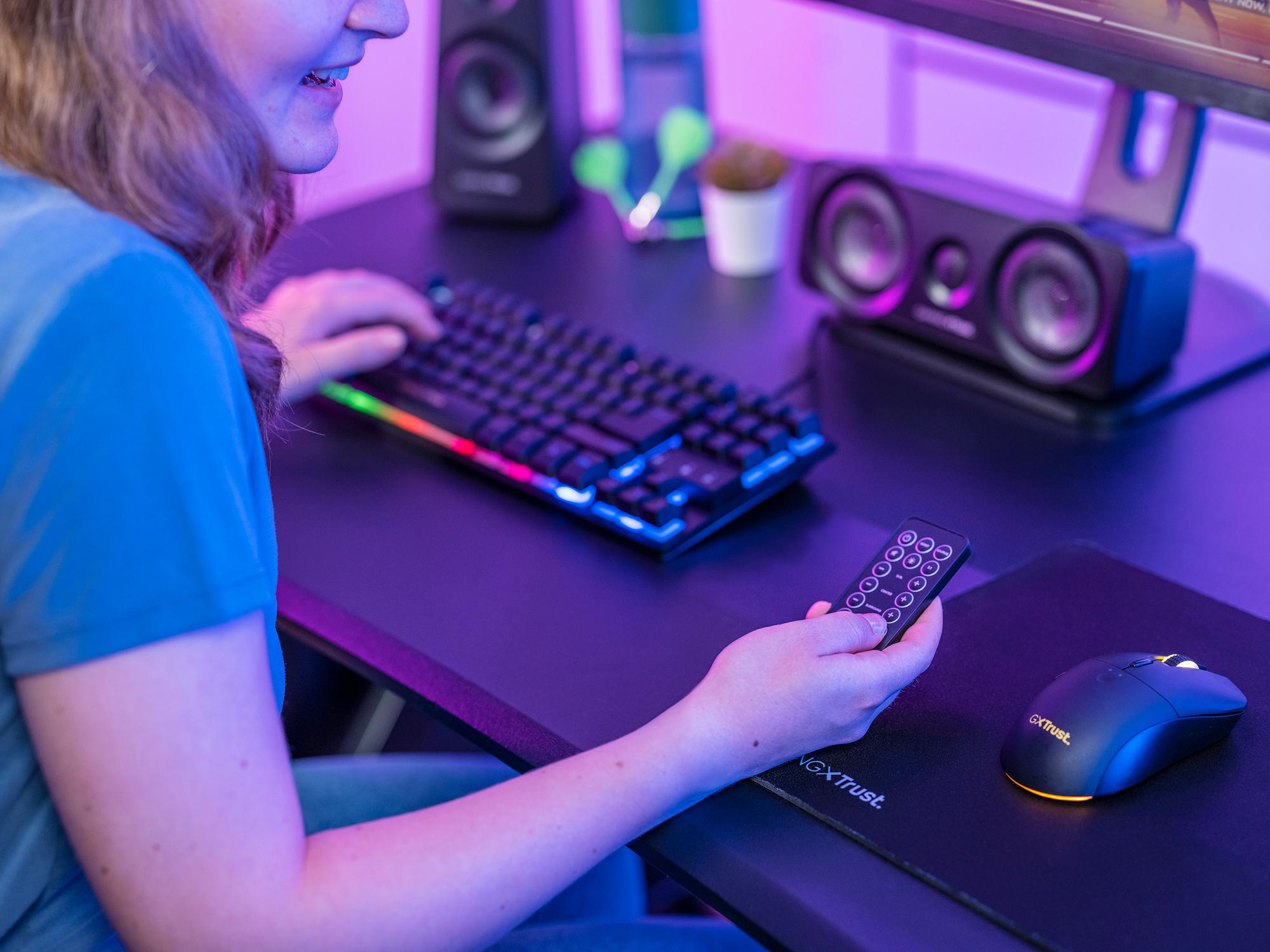 Eine Frau sitzt an einem Schreibtisch mit Gaming-Tastatur, Maus und Lautsprechern und hält eine Fernbedienung.