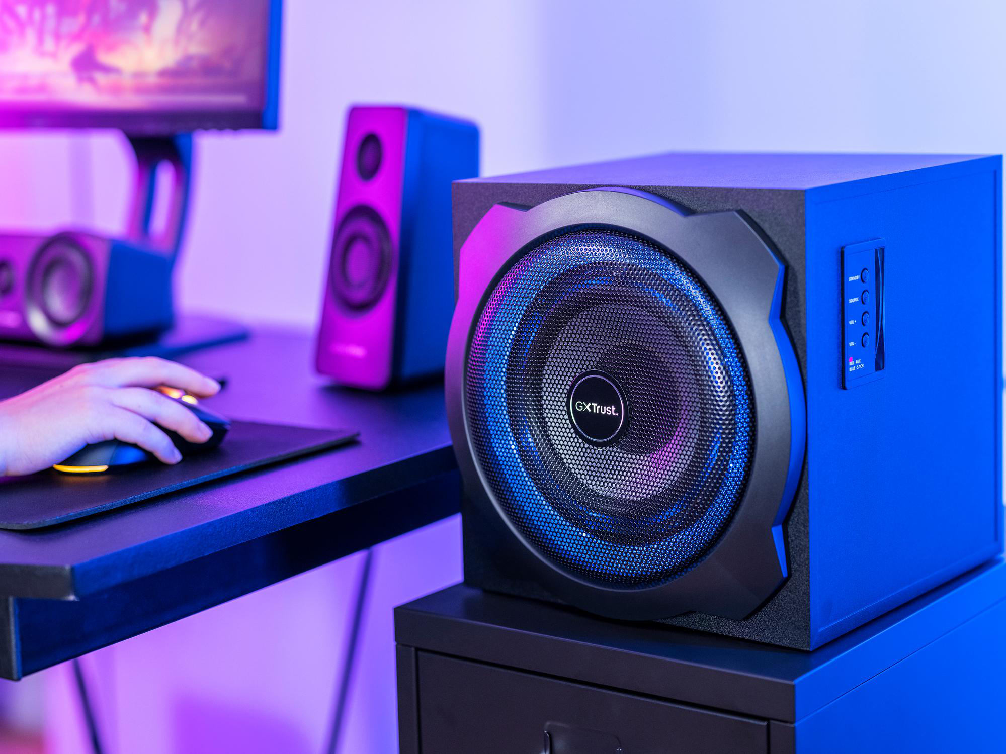 Ein schwarzer Subwoofer mit blauem und pinkem LED-Licht steht auf einem Schreibtisch neben einem Computer und Lautsprechern.