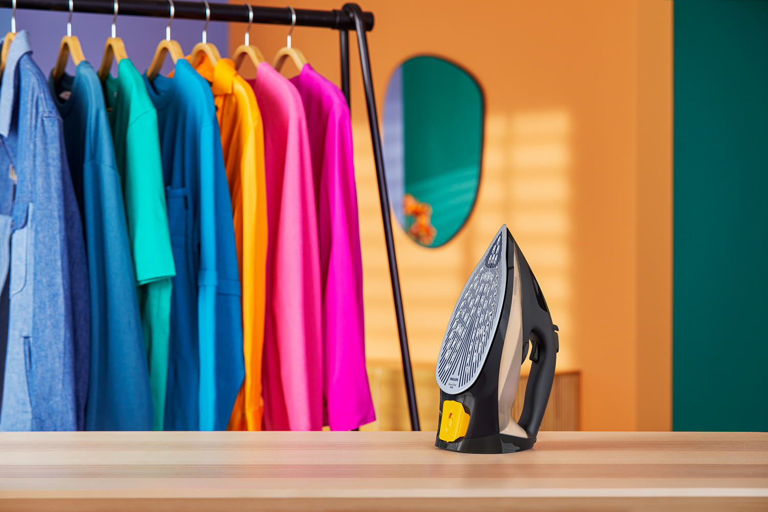 An iron on a wooden table, with a rack of colorful shirts in the background. Orange and green wall.