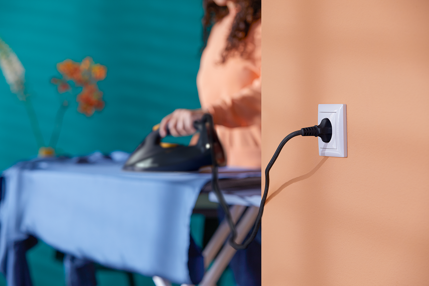 A woman ironing on a blue ironing board with a plug plugged into a wall outlet.
