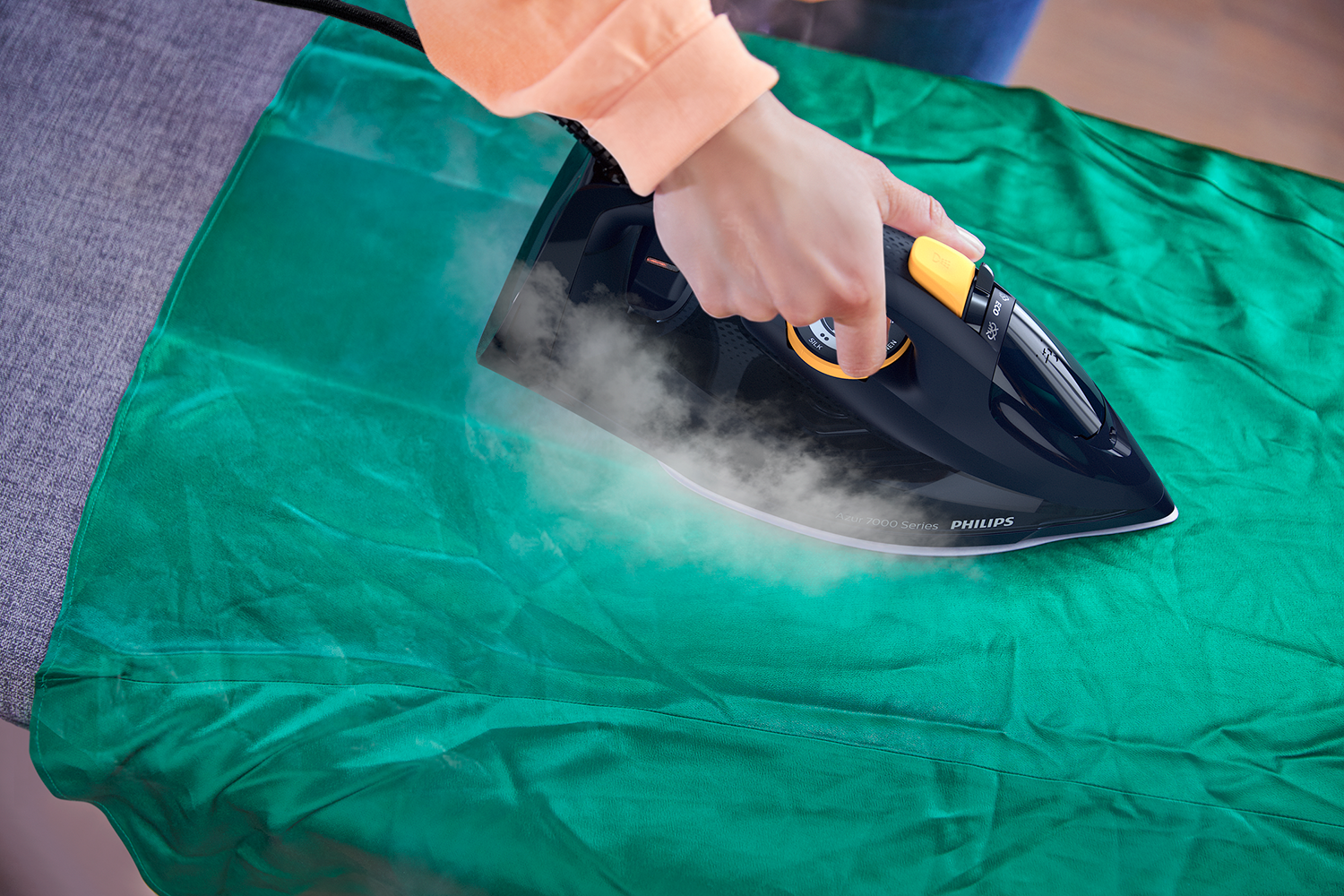 Person ironing green fabric, steam rising. Black iron, yellow dial. Grey ironing board. Bright, detailed shot.