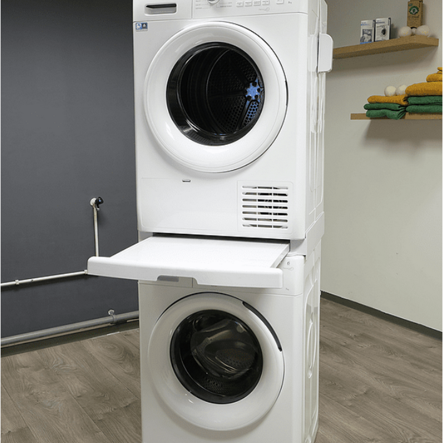 Stacked white washer and dryer. A tray is extended from the dryer. Grey wall, wooden floor.
