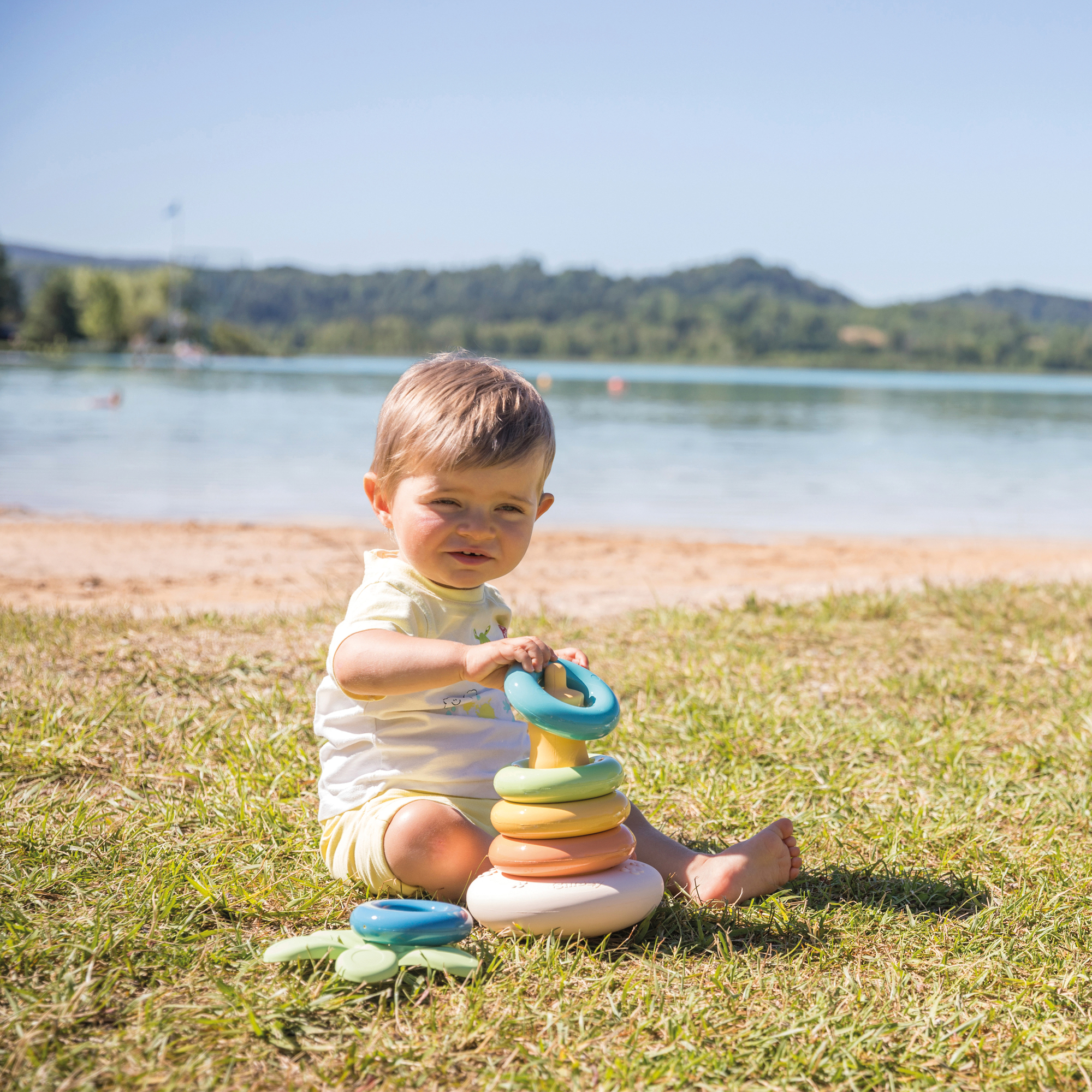 Ein Kleinkind, das auf Gras an einem See sitzt und mit einem Spielzeug-Ringstapler spielt. Blaue, grüne, orange und gelbe Ringe.