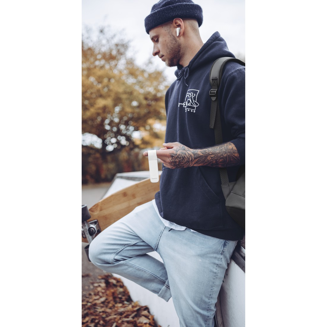 Ein Mann mit Mütze, hält ein kleines weißes Gerät, mit Tätowierungen und einem Skateboard. Draußen, Herbst.