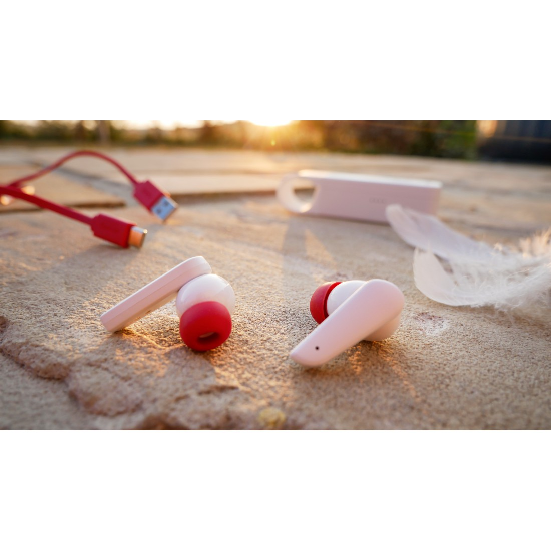 Paar rosa kabellose Ohrhörer mit roten Akzenten, die auf einer sandigen Oberfläche mit Feder und Kabel liegen.