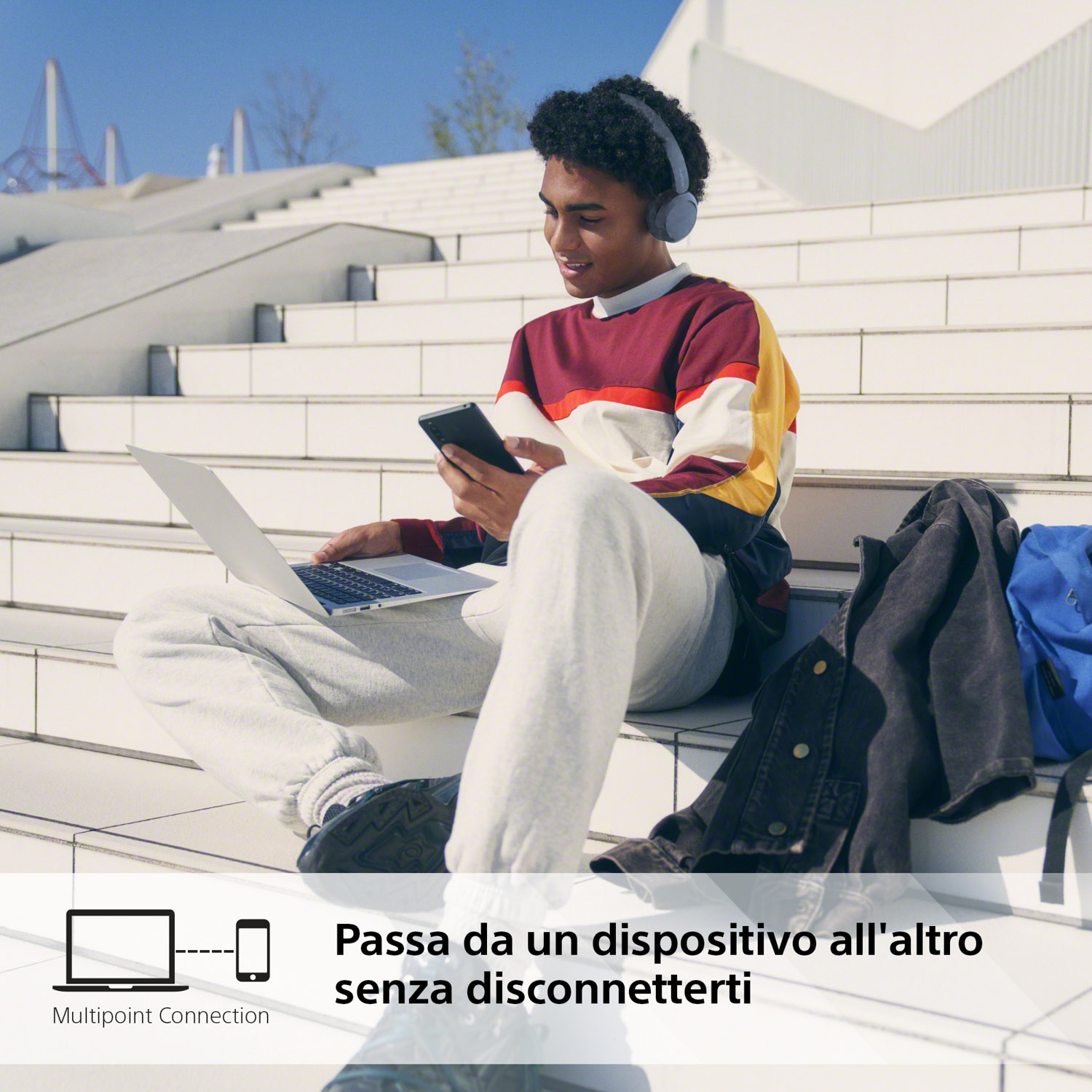 Giovane seduto sulle scale, che usa laptop e telefono, con cuffie.