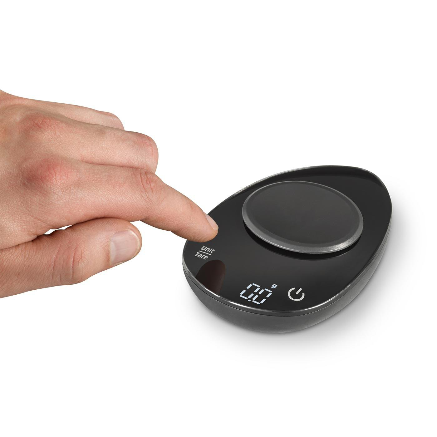 Eine schwarze Waage, eine Hand drückt die 'Unit/Tare'-Taste. Das Display zeigt 0,0.