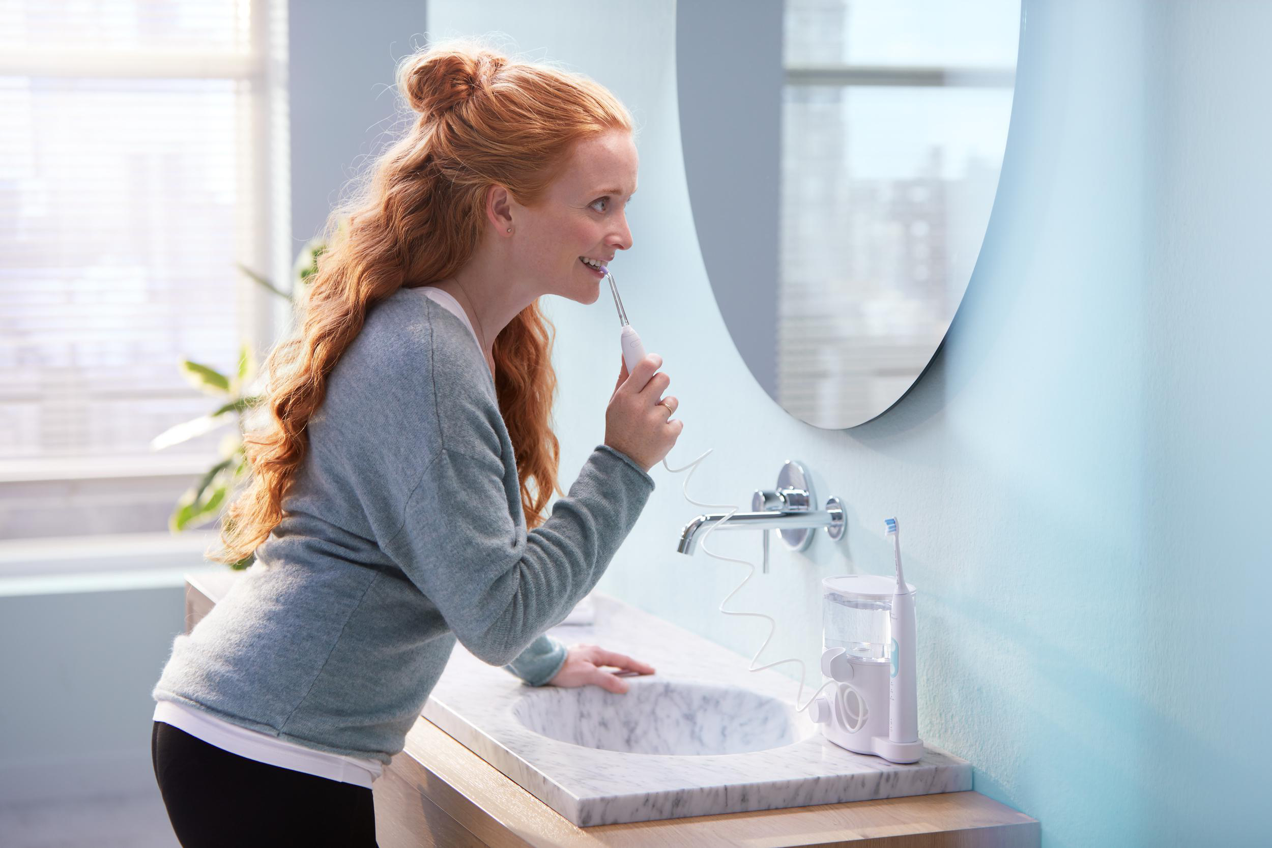 Eine Frau mit roten Haaren putzt sich im Badezimmer mit einem Wasserflosser die Zähne.