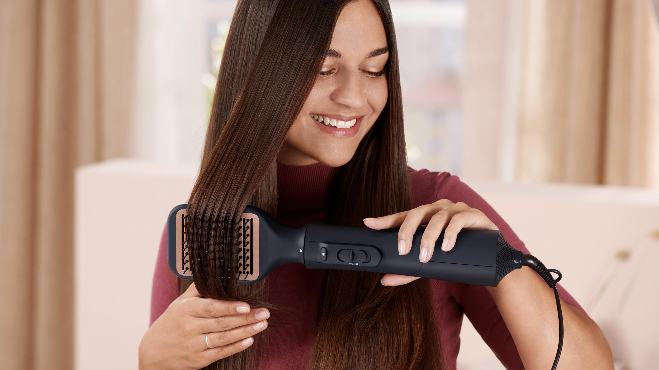 Eine Frau, die ihr Haar mit einem schwarzen Haarstyling-Werkzeug stylt, lächelt, drinnen.