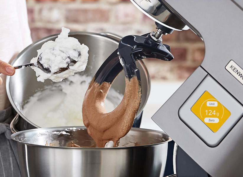 Ein Standmixer verrührt Teig, ein Löffel schöpft Teig aus einer Metallschüssel.