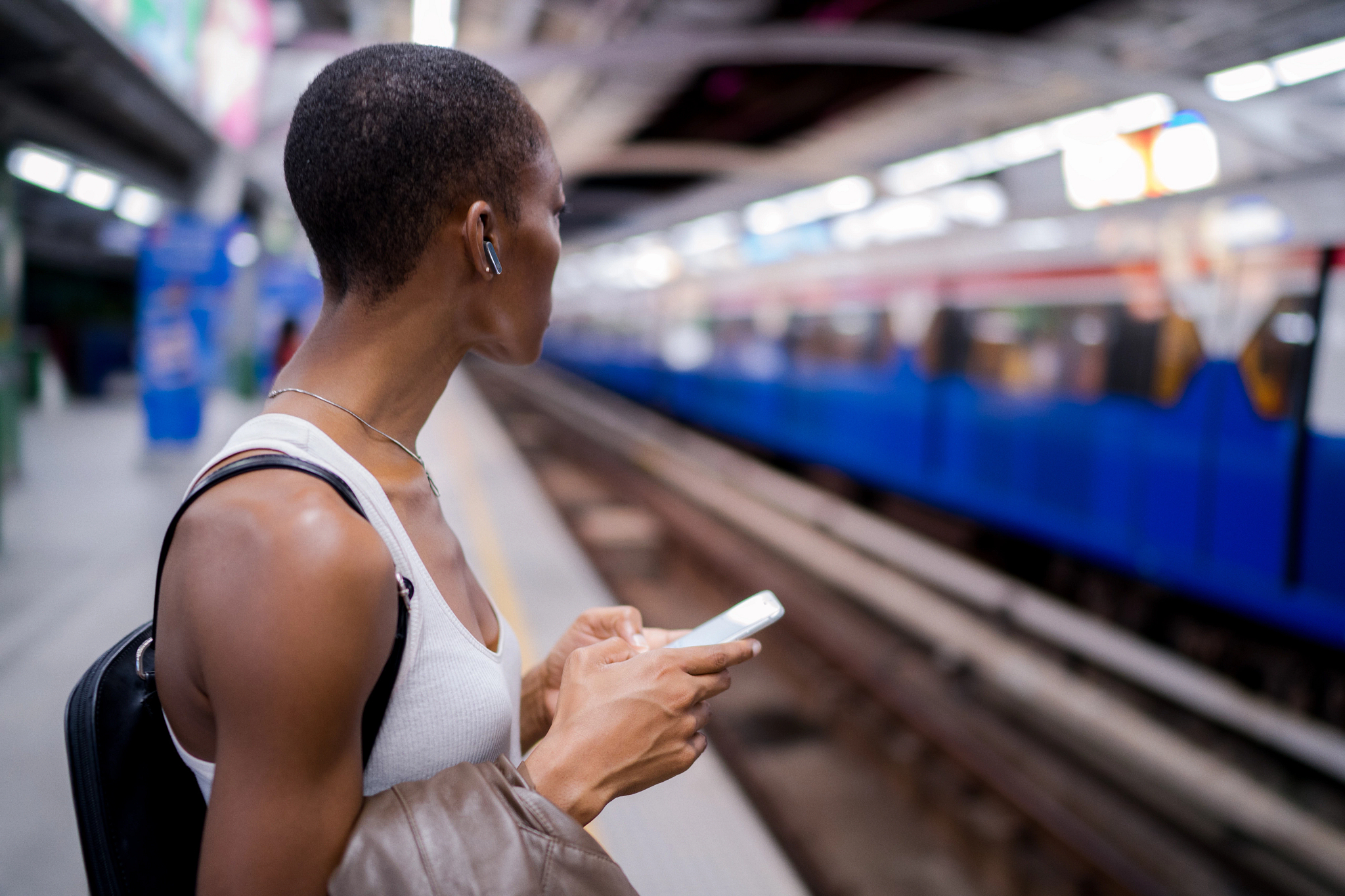 Frau am Bahnhof benutzt Telefon, mit Ohrhörern. Ein Zug ist im Hintergrund.