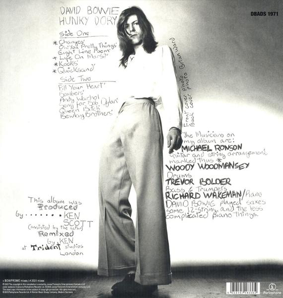 David Bowie, en chemise et pantalon blancs, debout avec une liste de titres de chansons sur un mur.
