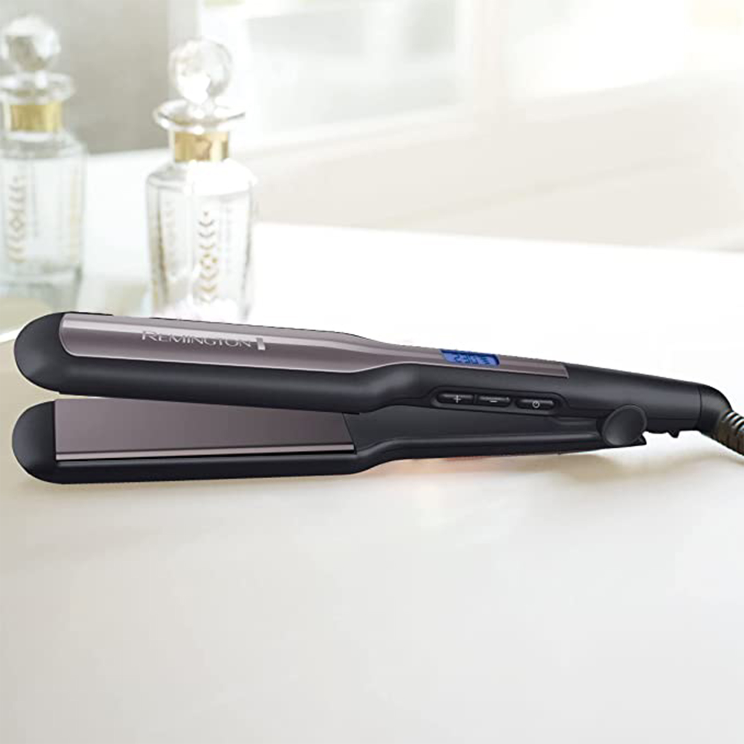 Piastra per capelli nera e grigia su una superficie bianca. Due bottiglie di profumo sullo sfondo.