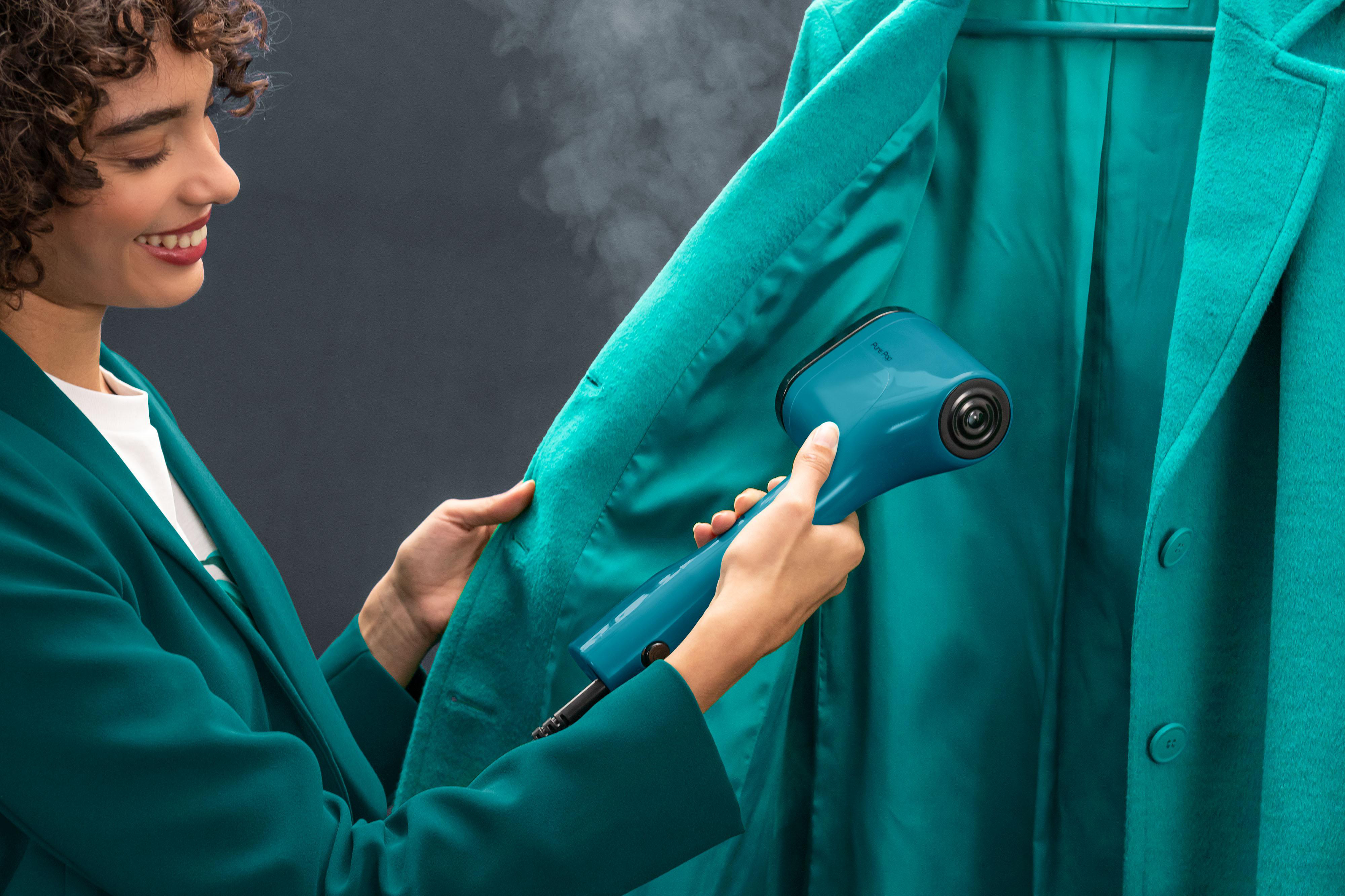 Frau, die einen tealfarbenen Mantel mit einem blauen Dampfgarer bedampft. Hintergrund ist eine dunkle Wand.