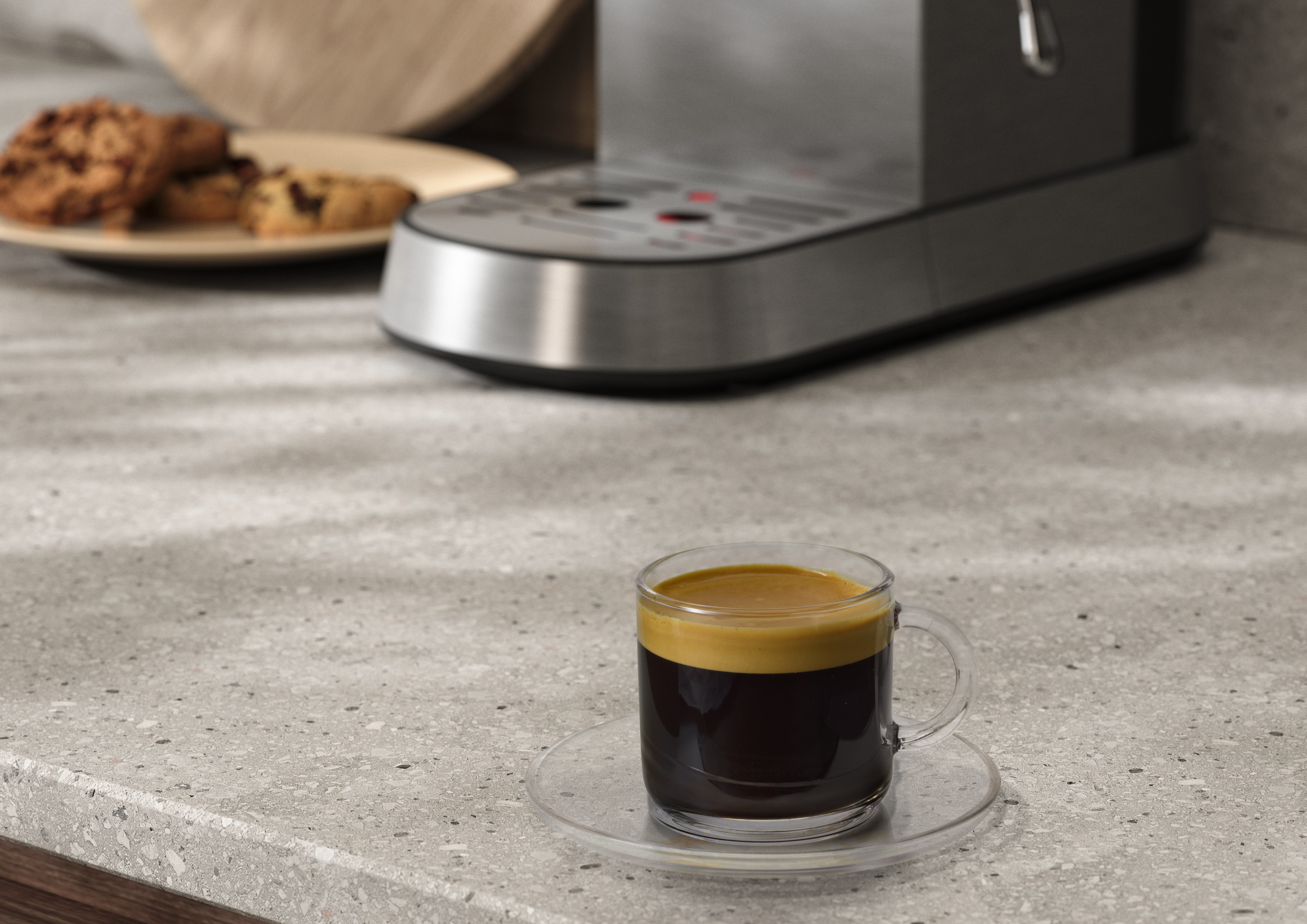 Gri bir tezgahta bir fincan espresso ve bir tabak kurabiye ve arka planda bir kahve makinesi.