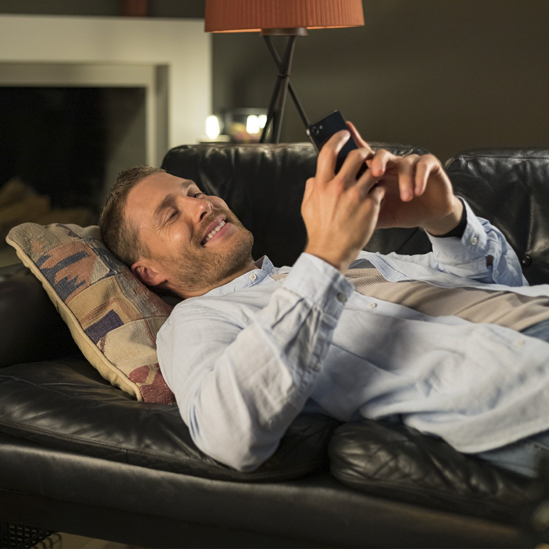 Mann liegt auf schwarzem Sofa, schaut mit Lächeln auf ein Telefon.