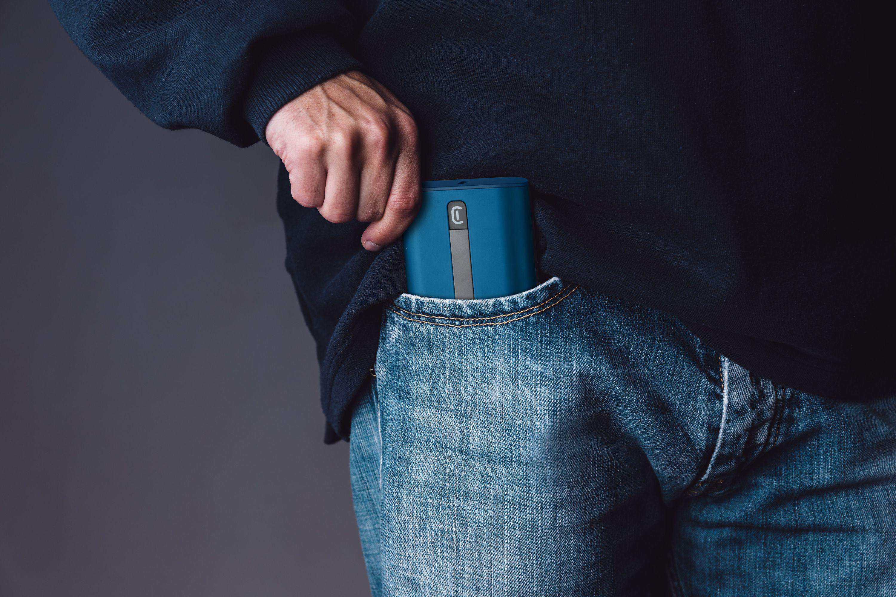 Eine Hand, die ein blaues Gerät in eine blaue Jeans-Tasche steckt. Dunkler Hintergrund.