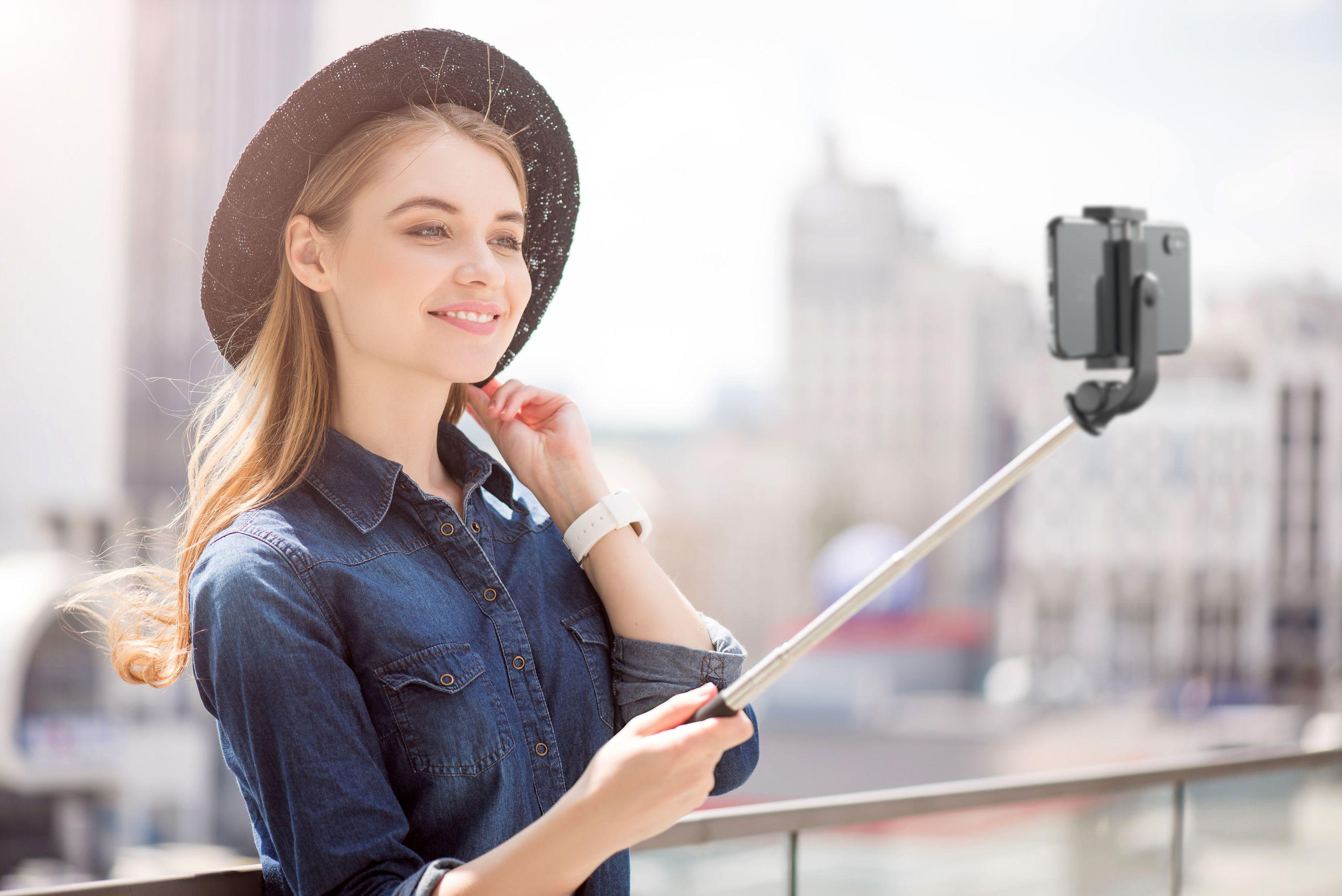 Eine junge Frau macht ein Selfie auf einem Dach mit Stadtblick im Hintergrund.