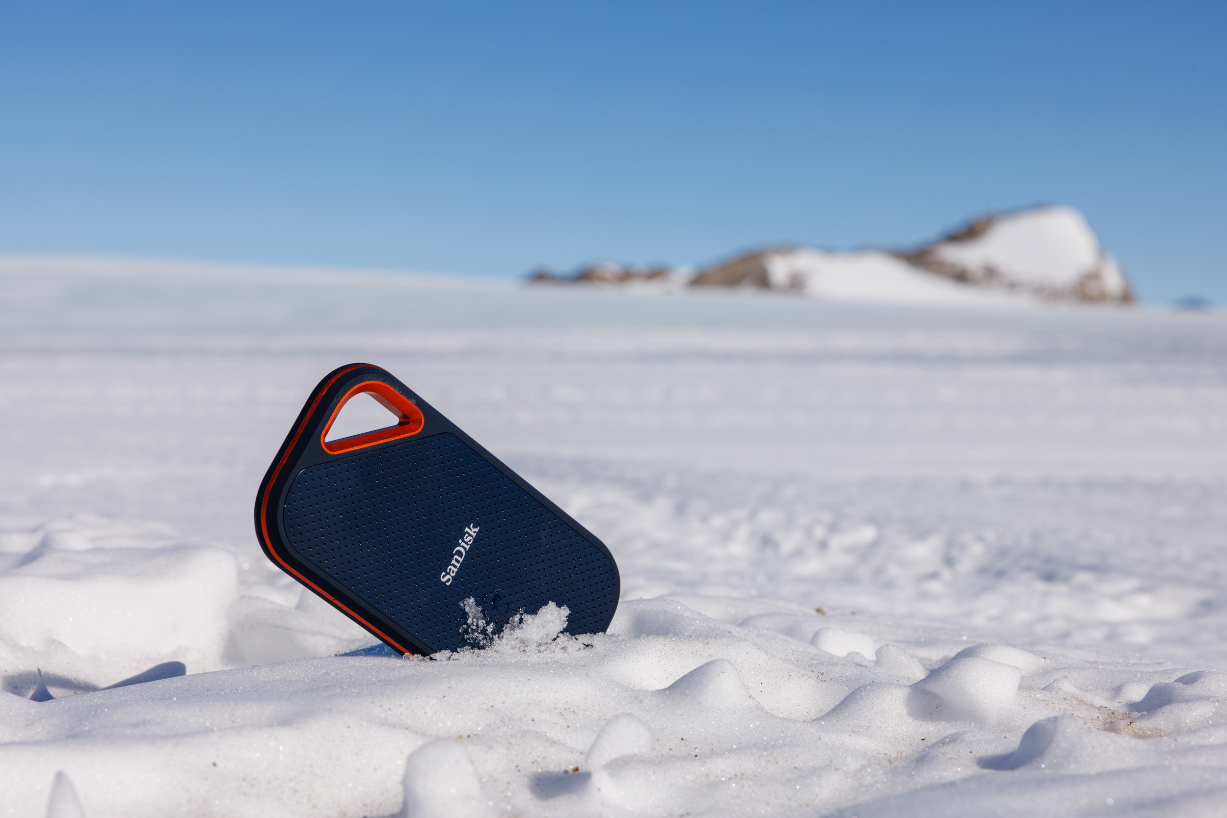 Kar üzerinde mavi ve turuncu Sandisk taşınabilir sabit disk. Arka planda mavi gökyüzü.