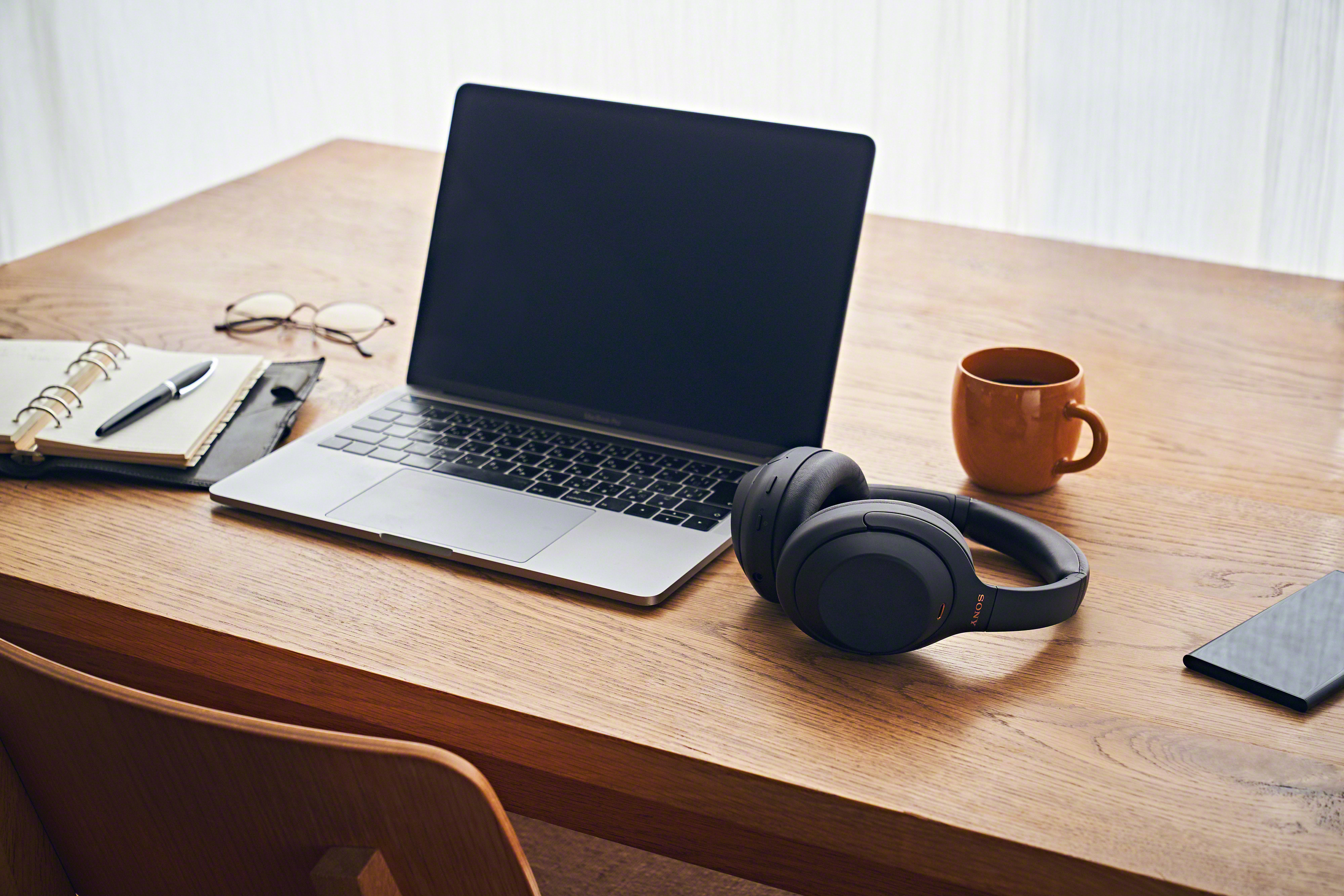 Laptop, Kopfhörer, Becher und Notizbuch auf einem Holztisch. Brille und Telefon sind ebenfalls vorhanden.