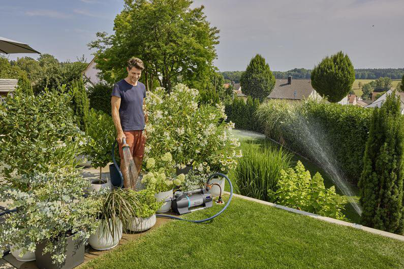 Mann wässert Garten mit Schlauch, daneben Gartengerät und Sprinkler.