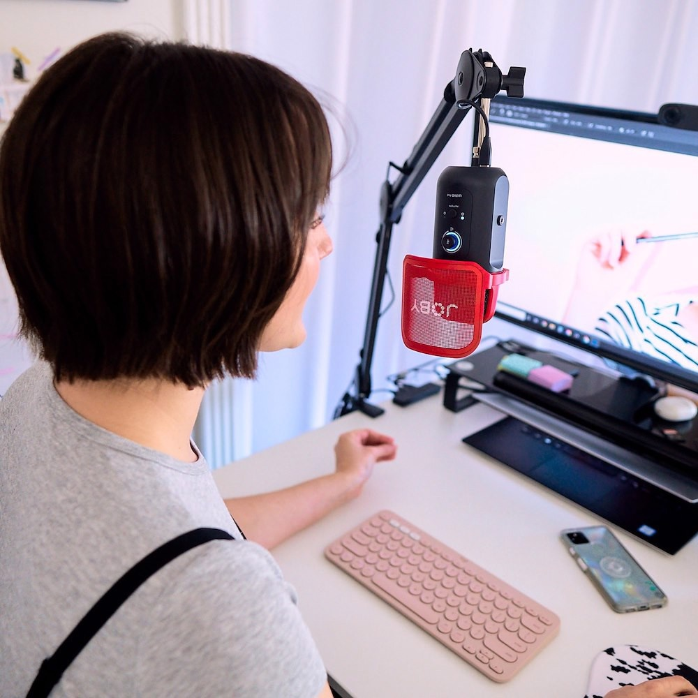 Frau hinter Schreibtisch mit Mikrofon, Monitor, Tastatur und Telefon. Der Hintergrund ist ein Raum.