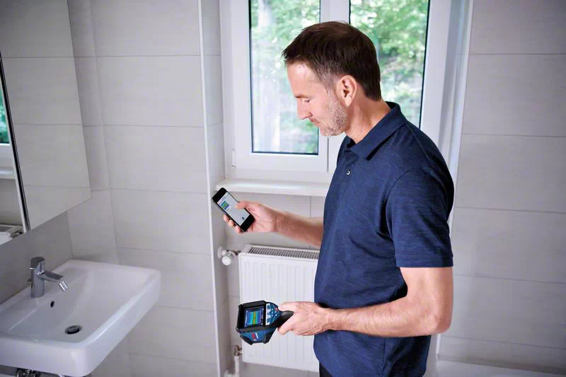 Egy férfi a fürdőszobában, kezében egy telefonnal és egy hőkamerával. Egy mosdókagyló előtt áll.