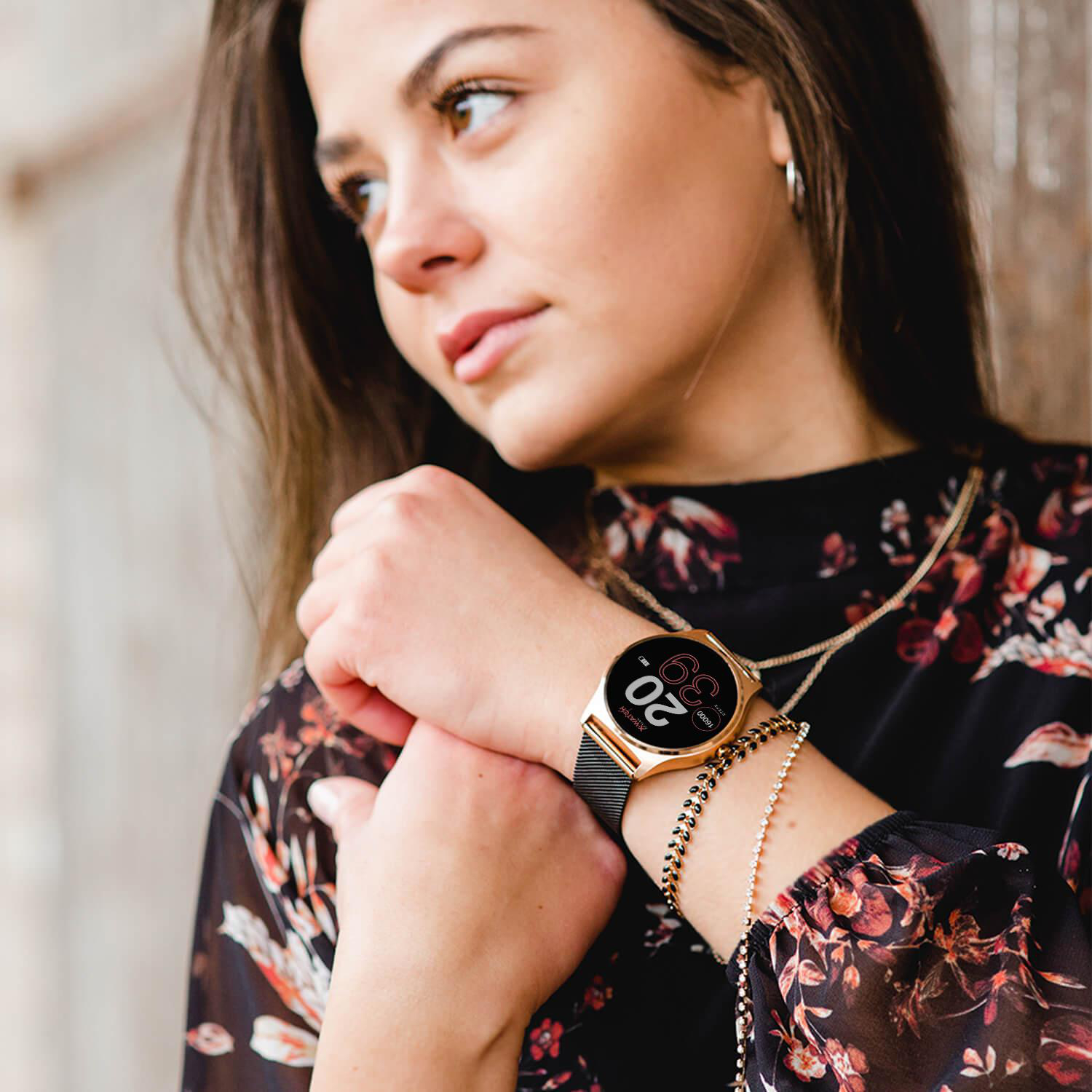 Eine Frau mit einer Uhr am Handgelenk, mit einem gemusterten Oberteil und Goldschmuck.