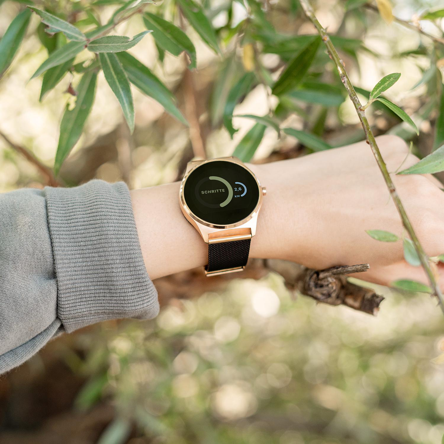 Eine Person trägt eine roségoldene Smartwatch, im Freien mit grünem Laub im Hintergrund.