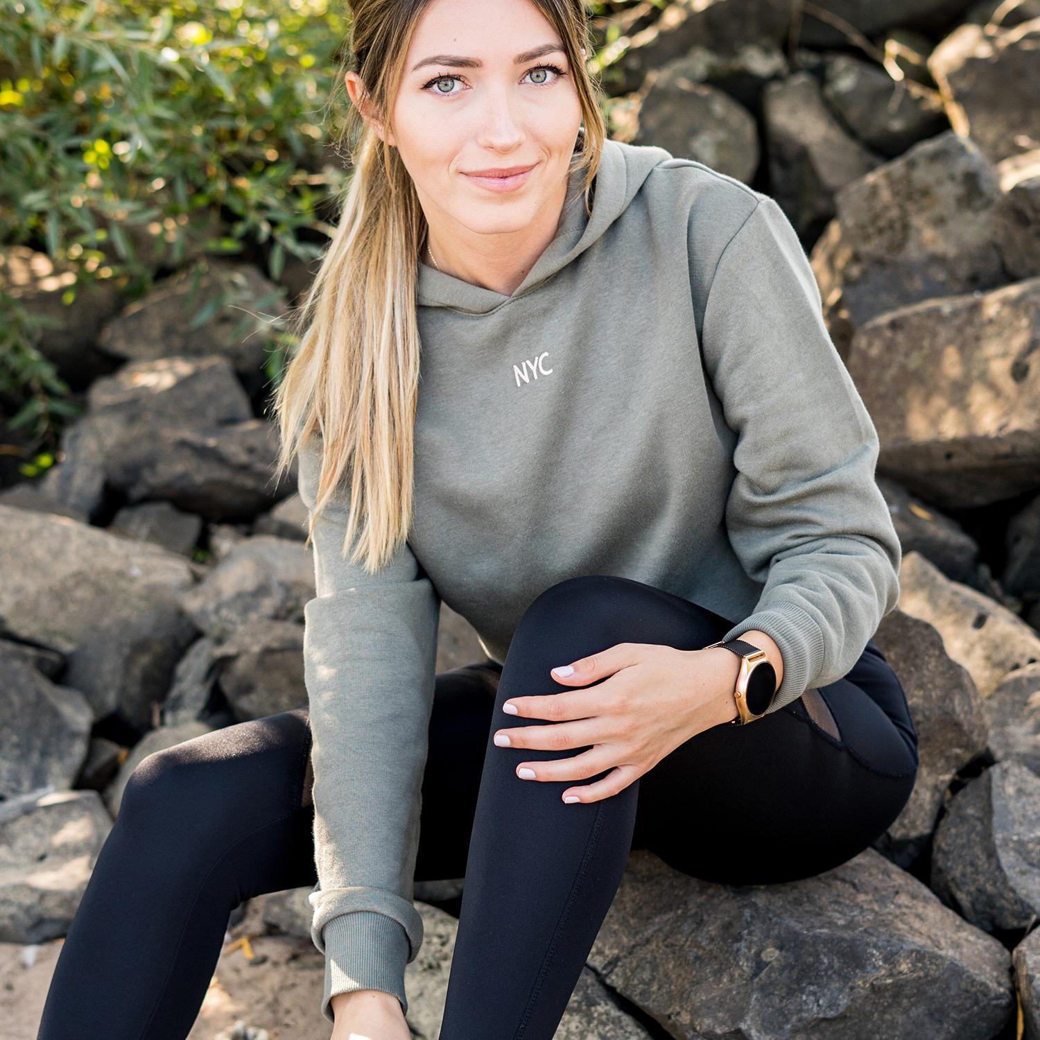Eine Frau in einem grünen Kapuzenpullover und schwarzen Leggings sitzt auf Felsen und lächelt in die Kamera.