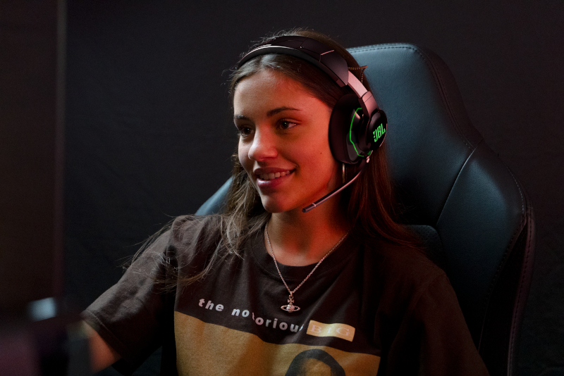 Eine junge Frau mit Headset lächelt. Sie sitzt in einem Gaming-Stuhl mit dunklem Hintergrund.