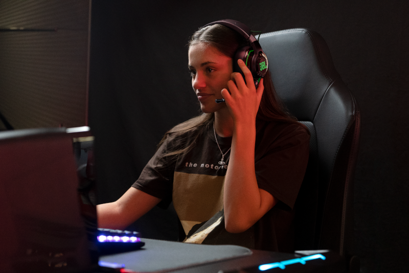Frau mit Kopfhörern und Mikrofon, die auf einen Monitor schaut und ein Spiel spielt. Die Kopfhörer haben ein JBL-Logo.