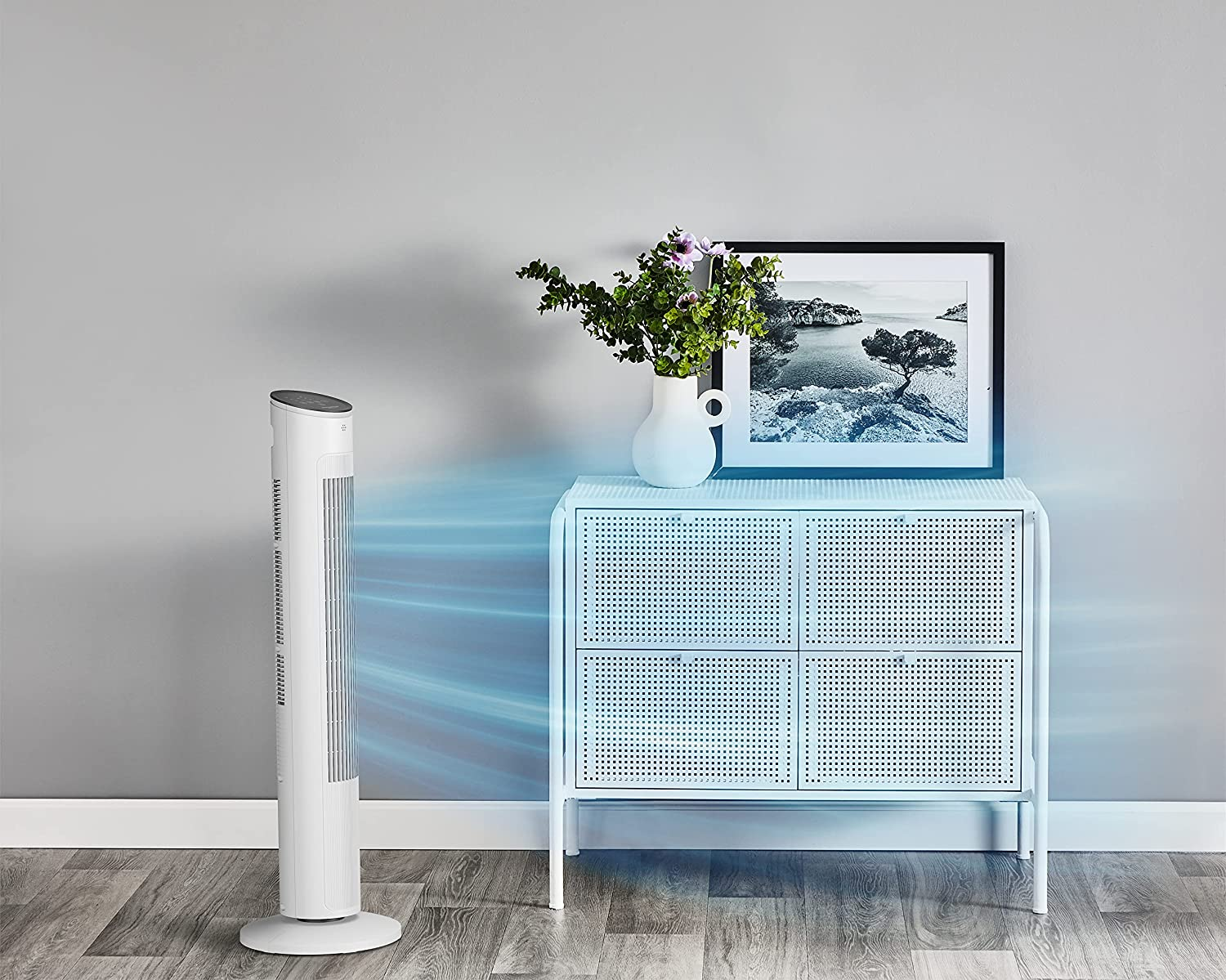 Un ventilatore a torre bianco soffia aria blu verso un mobile bianco con vaso e quadro.